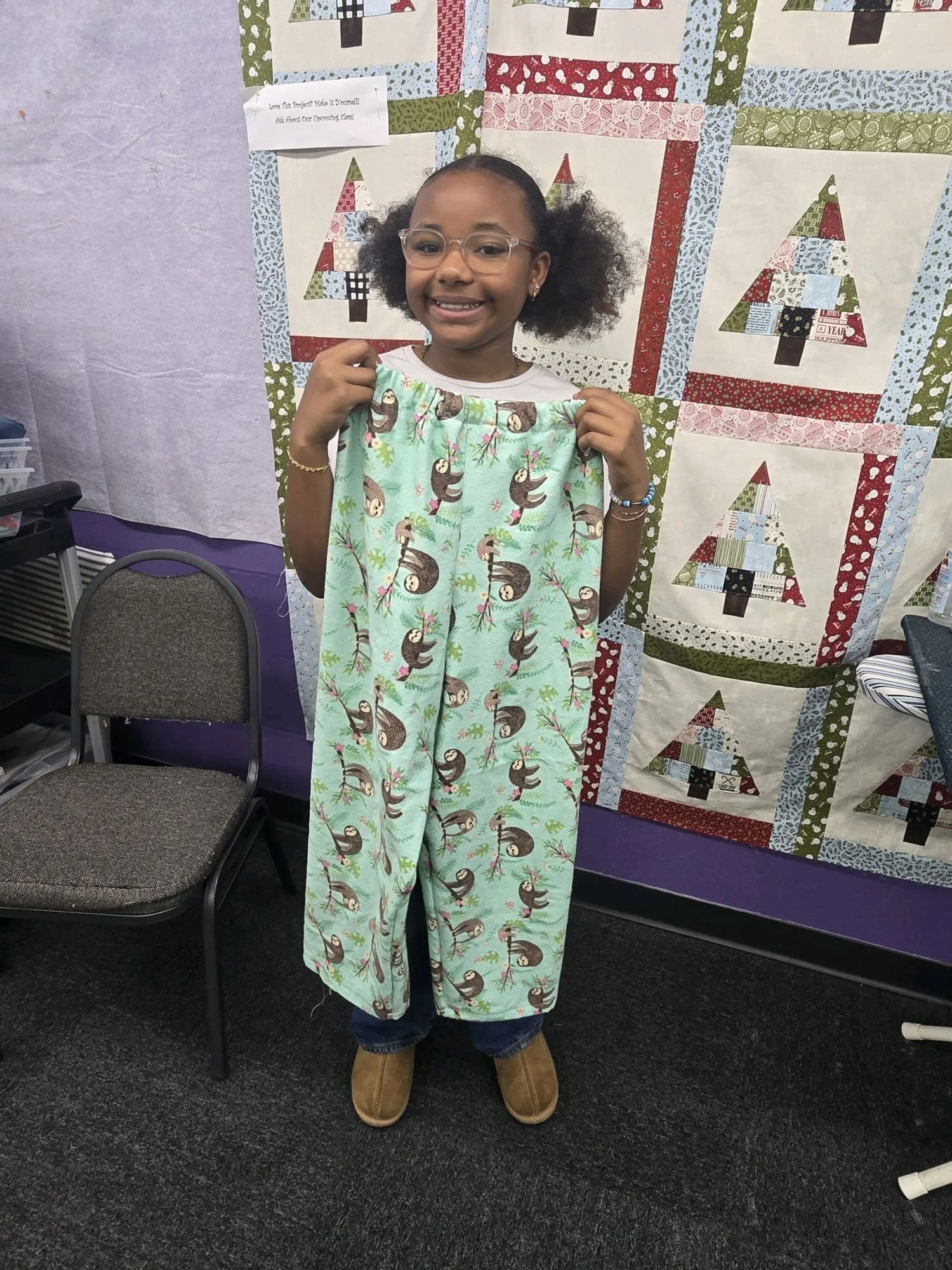 A young girl with glasses smiling and holding up a pair of pajama pants with a sloth pattern, standing in front of a holiday-themed quilt display.
