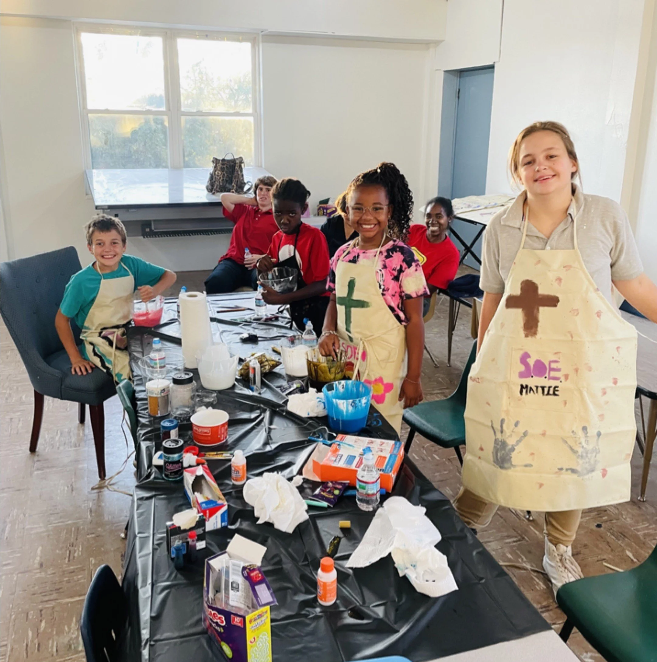 A group of children in aprons participating in an arts and crafts activity around a table covered with supplies, with some children smiling and others engaged, in a room with large windows and a piano.