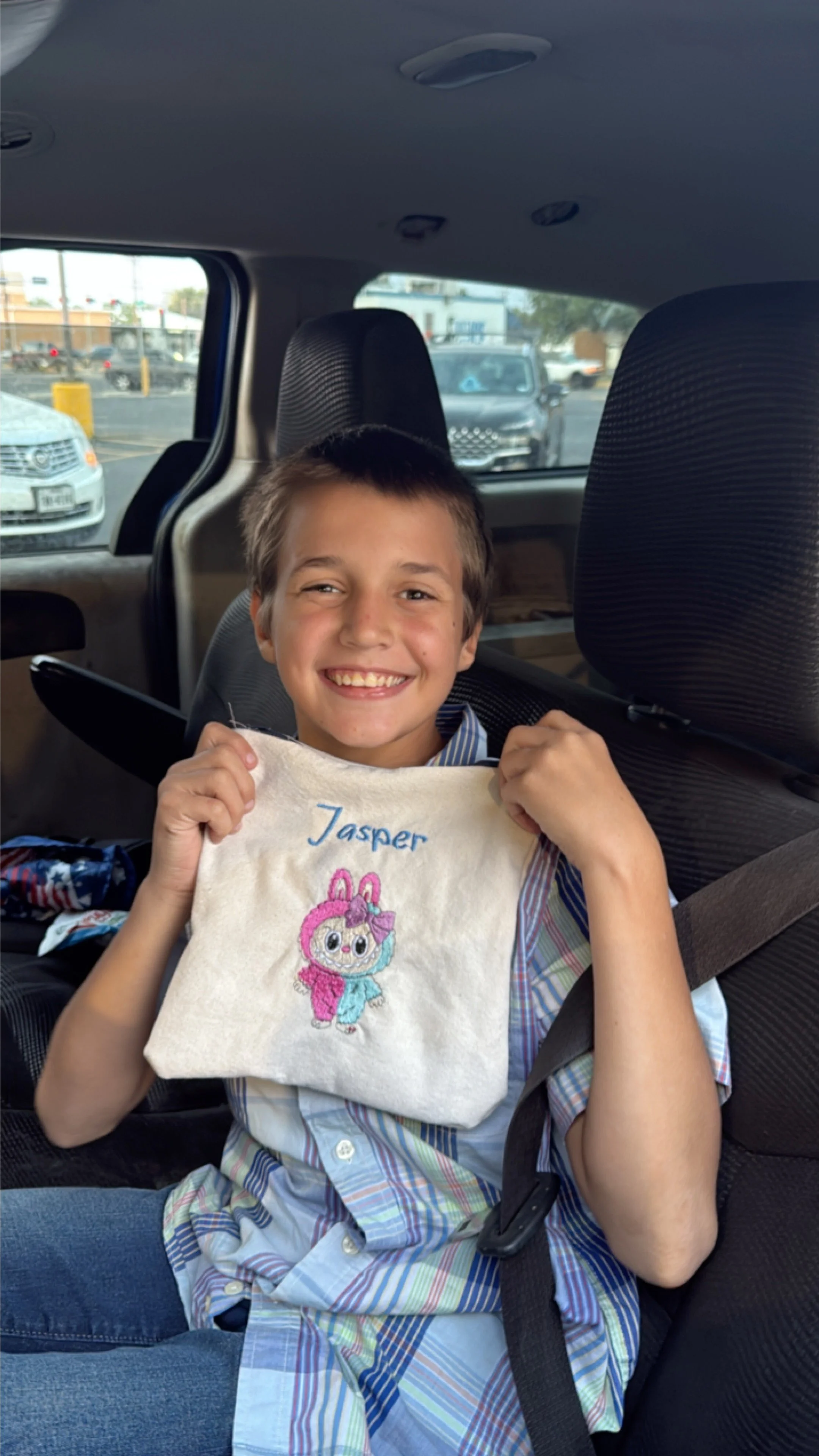 A smiling young boy sitting in the back seat of a vehicle, holding a embroidered bag with the name 'Jasper' and a cartoon bunny character. The boy is wearing a colorful, striped shirt and appears happy.