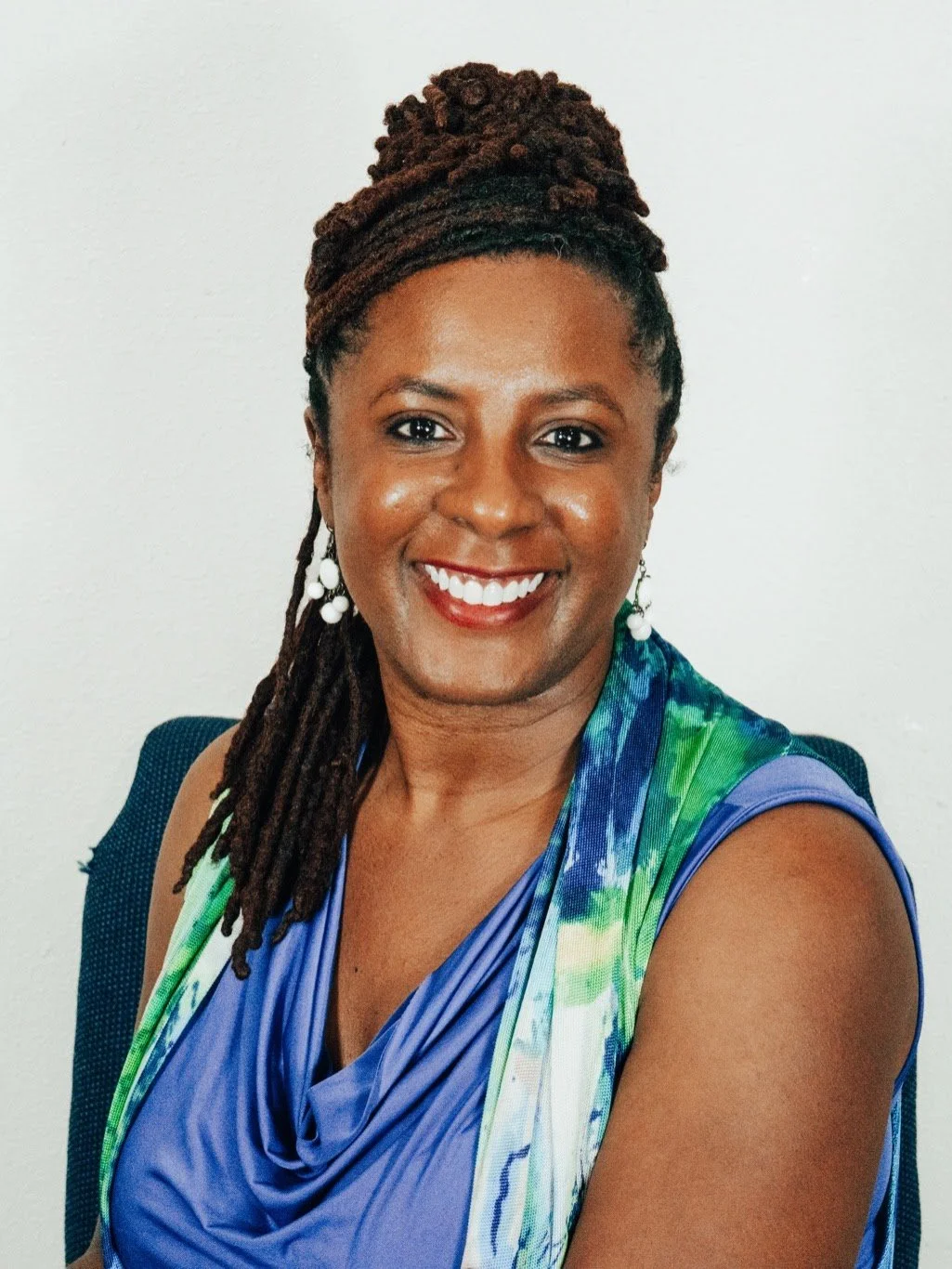 A woman with brown skin, styled in dreadlocks tied up with a headband, smiling at the camera. She is wearing earrings and a colorful silk or satin sleeveless dress.