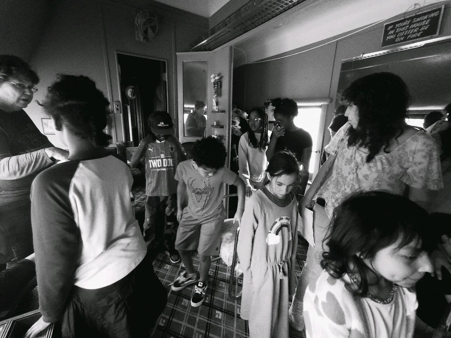A group of children and adults crowded inside a room, some standing and some sitting. The room has patterned carpet, a door, and a sign that reads 'If you're standing in this house you better be on fire.'