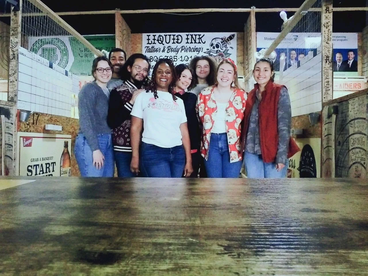 Group of eight smiling people standing inside a bar or restaurant with wooden walls and plywood. In the background, there's a sign for a tattoo shop called 'Liquid Ink' and various posters and signs on the walls.