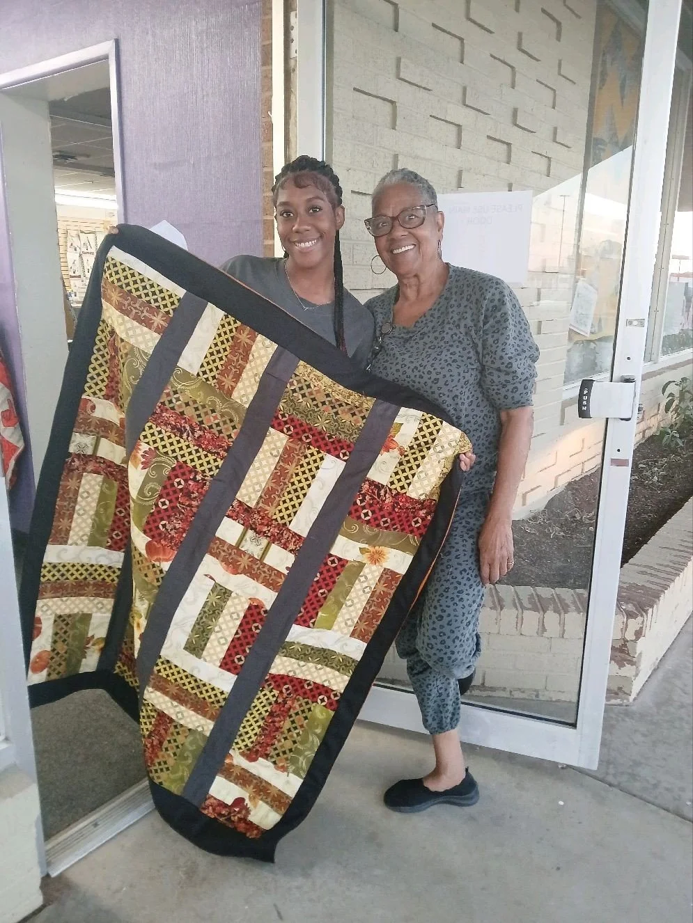 Two women standing outside a building, smiling, holding a colorful quilt with African-inspired patterns. The older woman has short gray hair and glasses, wearing a gray patterned outfit. The younger woman has braided hair, wearing a dark shirt.