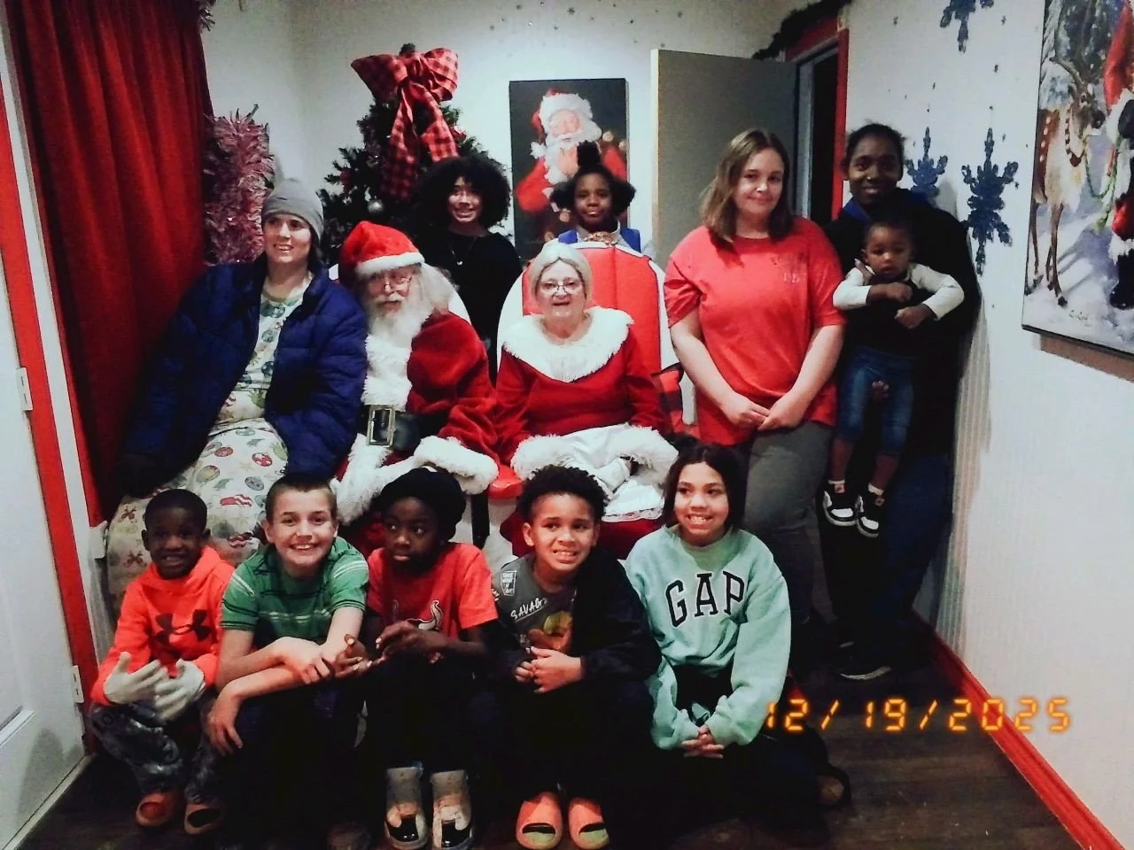 Group of children and adults, including Santa Claus and Mrs. Claus, gathered in front of a decorated Christmas tree. The children are seated and standing, some smiling, in a festive indoor setting.