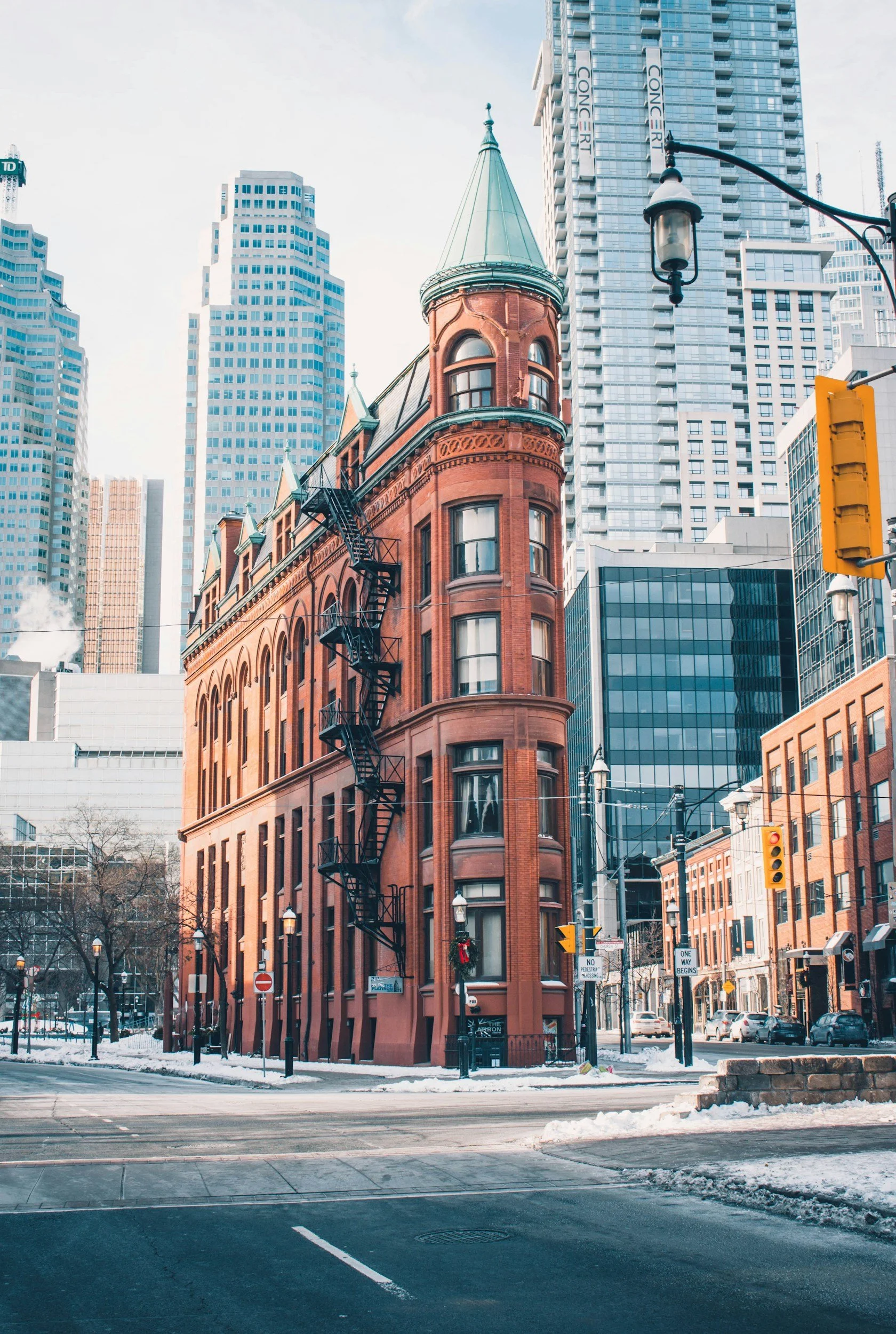 A historic red brick building with a turret and fire escape, surrounded by modern skyscrapers in an urban cityscape with snow on the ground.