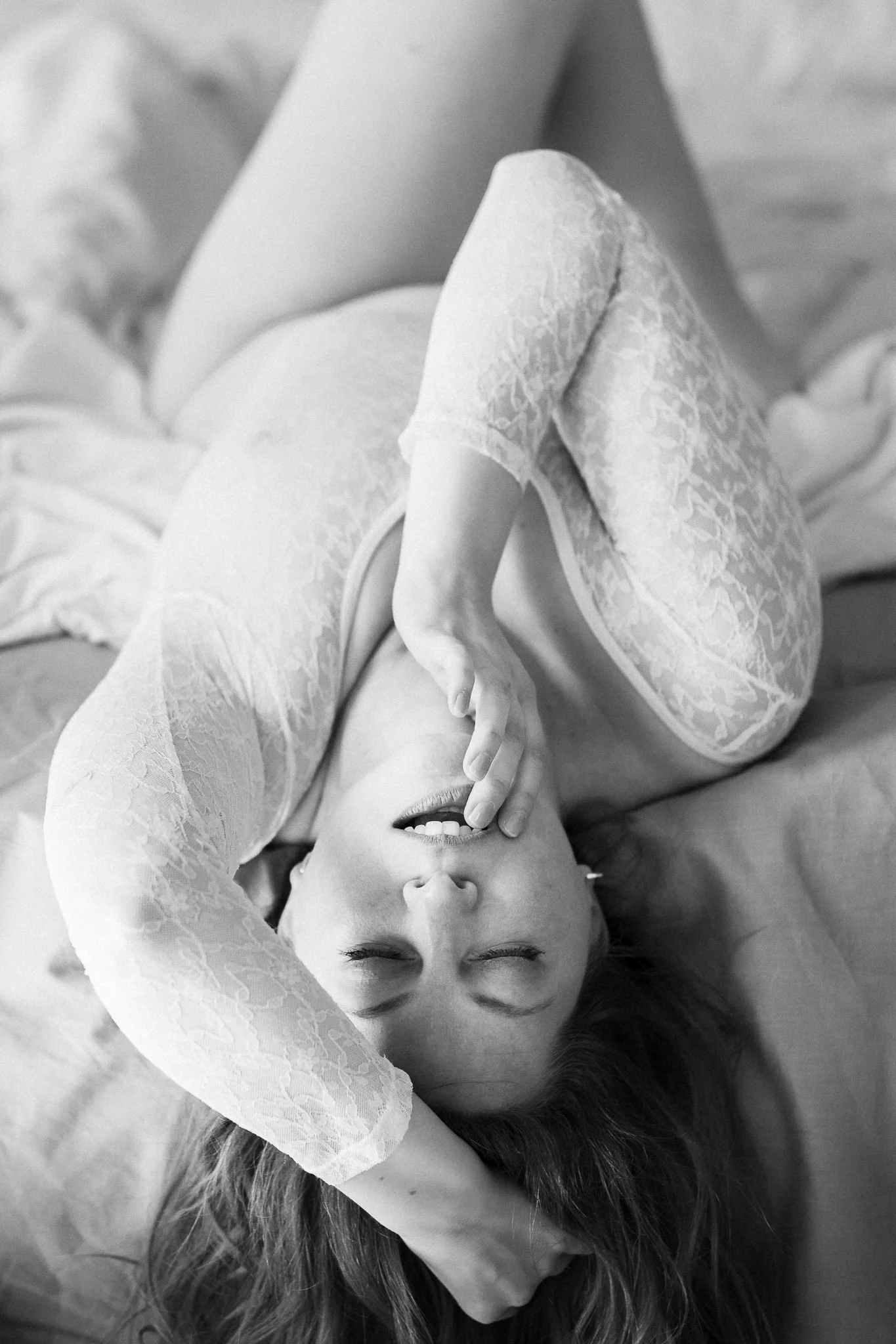 A woman lying down with her hand on her face, wearing lace clothing, smiling with her eyes closed. Her hair is observed around her head and a large flower is near her face.