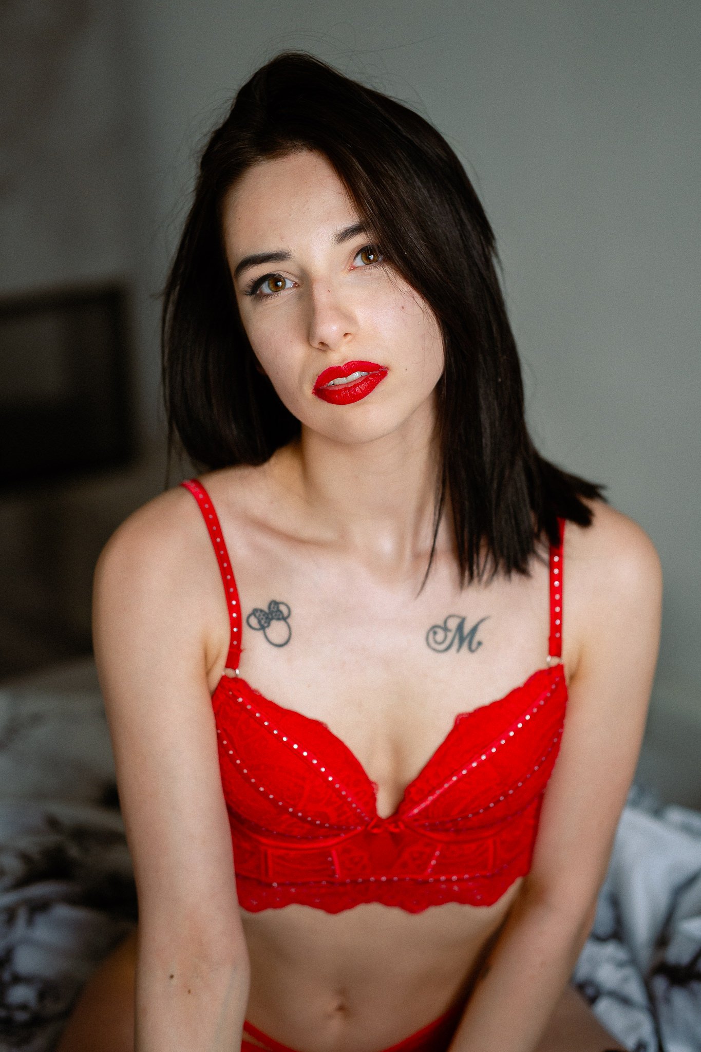 A woman with dark hair and red lipstick wearing a red lace lingerie top with polka dot straps, displaying tattoos on her chest.