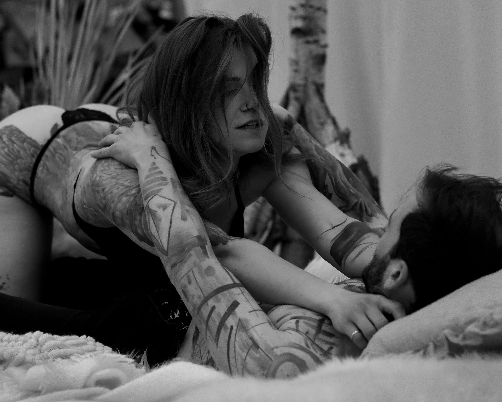 A woman with tattoos and a nose ring lays on her stomach on a bed, resting her head on her crossed arms, with her hair falling over part of her face.
