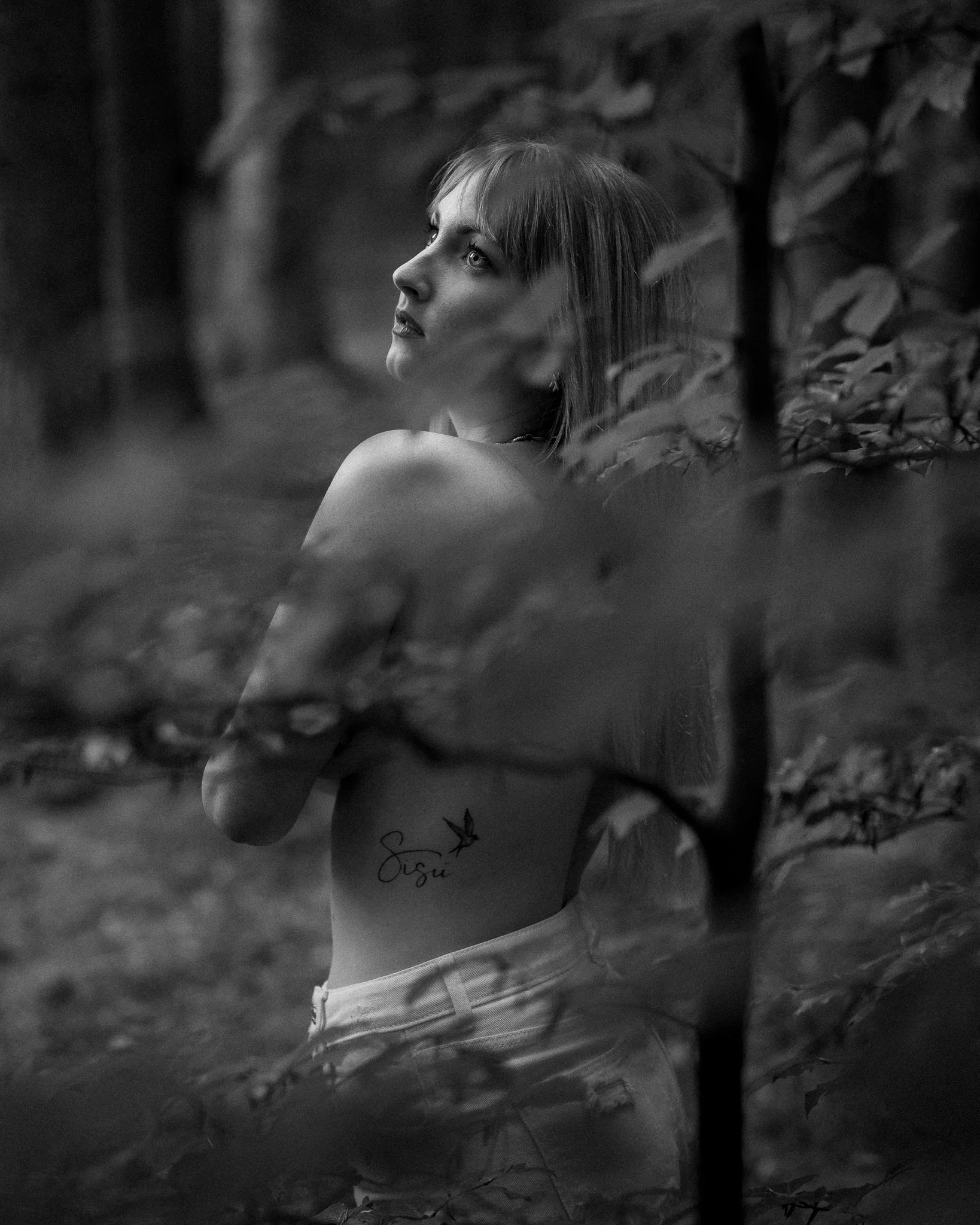 A black and white photo of a woman with light-colored hair and tattoos on her arm, sitting outdoors among foliage. She is looking upwards with a thoughtful expression.