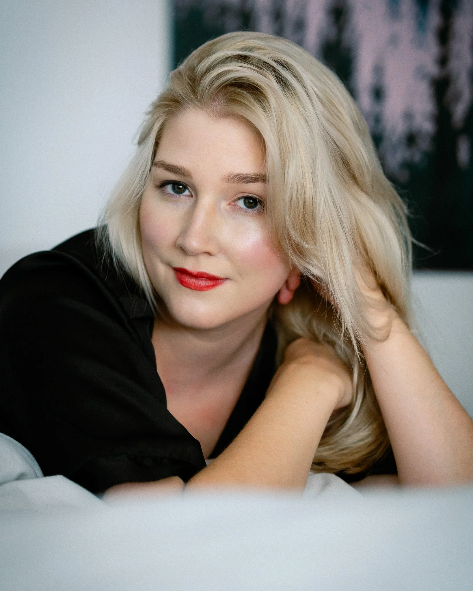 A woman with blonde hair and blue eyes resting her face on her hand, looking at the camera, with a dark patterned background.