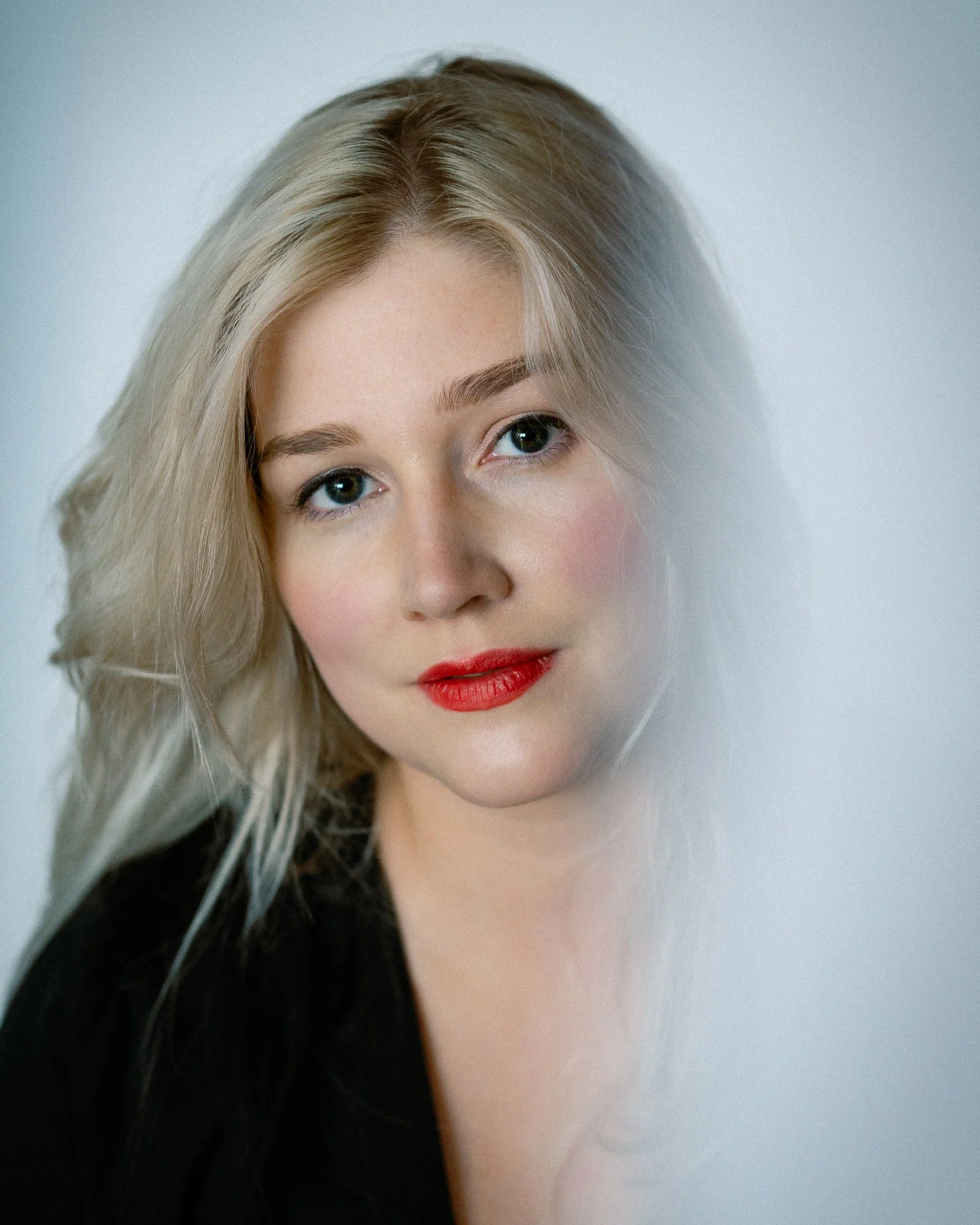 Partial view of a woman's face with blonde hair, bright eyes, red lipstick, against a plain light background.