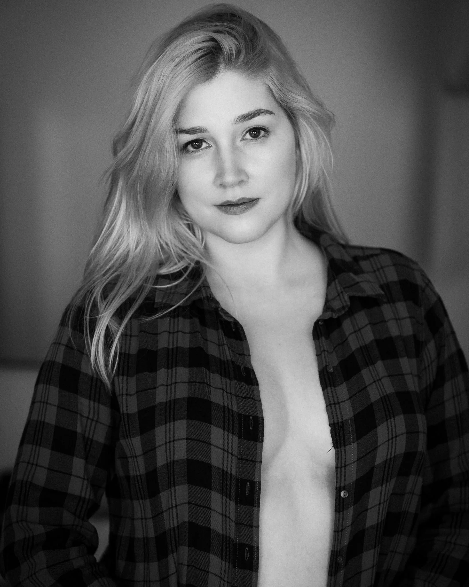 A woman with long, wavy hair wearing a checkered shirt, with part of her chest visible, smiling softly. Black and white photo.