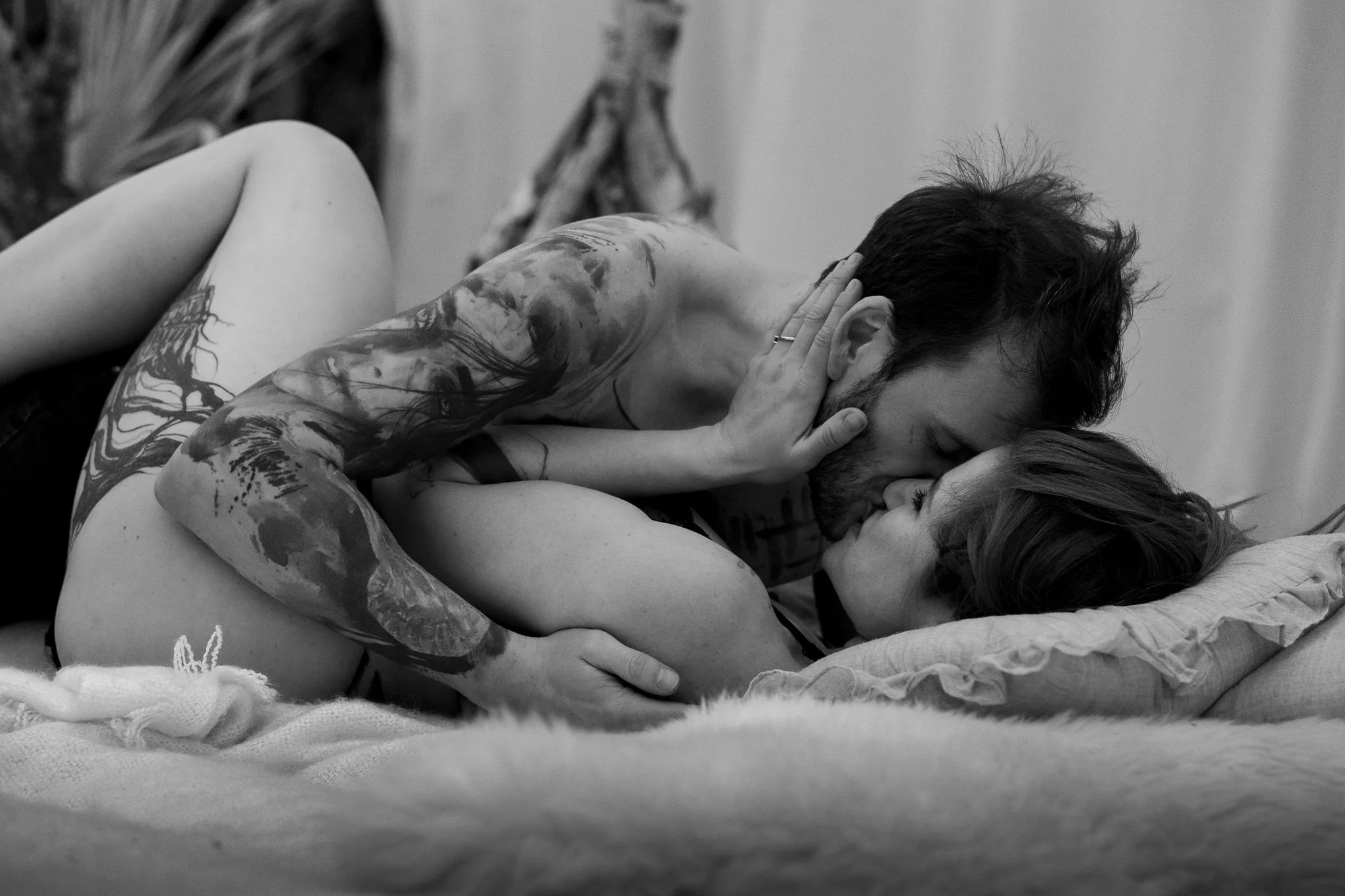 A couple lying on a bed, engaged in a passionate kiss, with the man shirtless and the woman with her arm around his neck, displaying tattoos on his arm and shoulder.