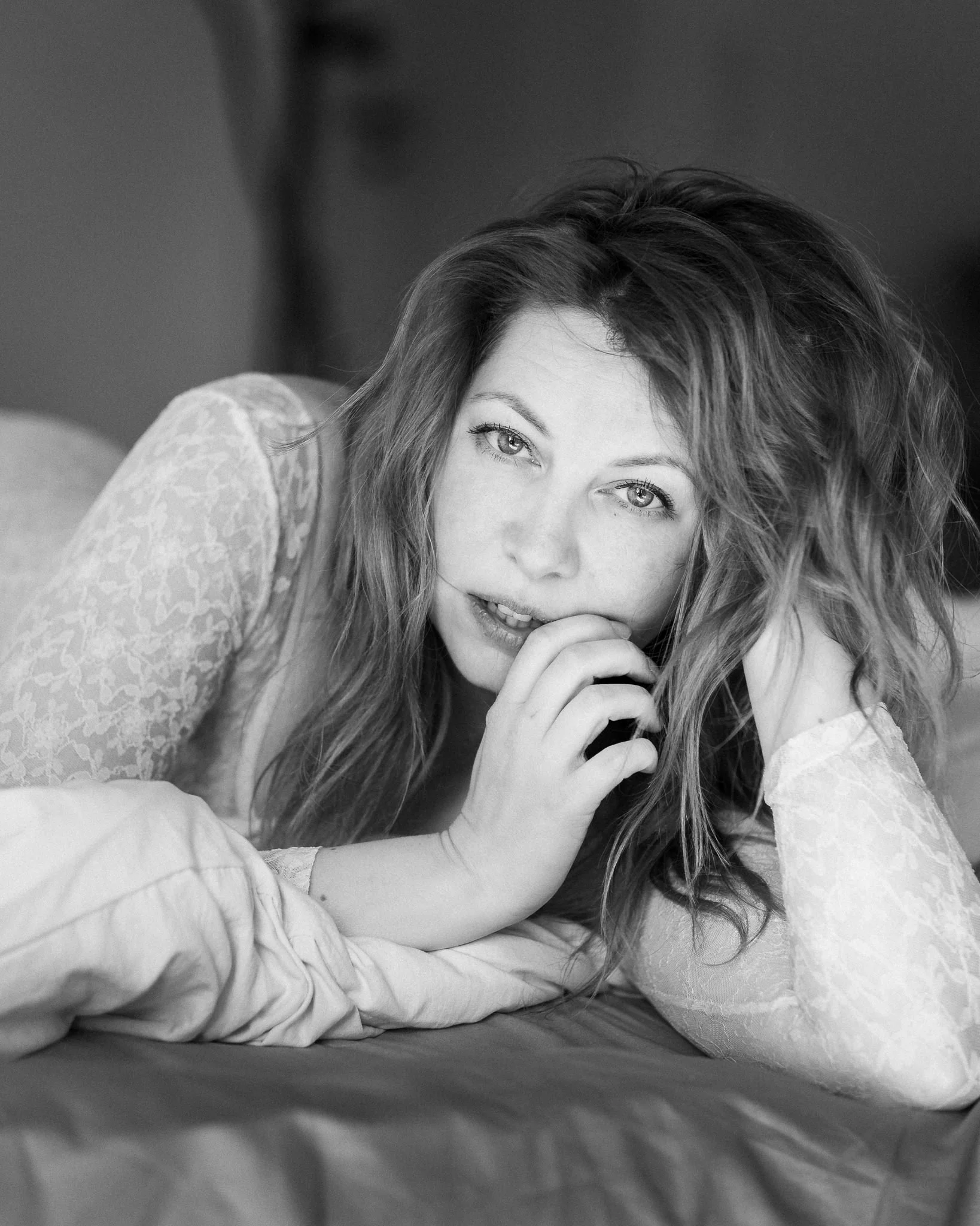 A black and white close-up photograph of a woman with long hair, lying on a bed, with her hand touching her lips, looking directly at the camera.