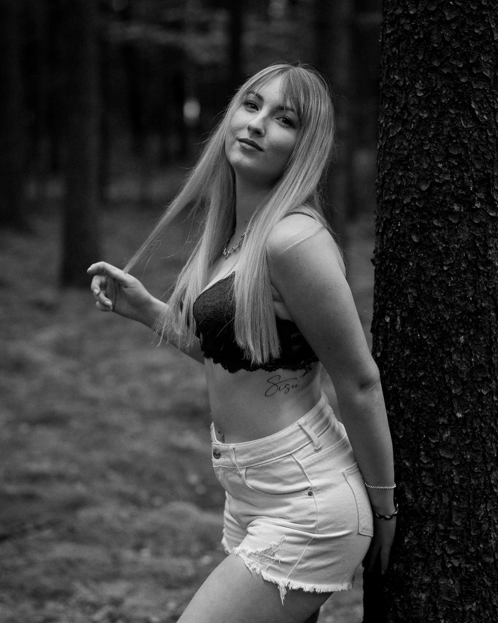 A young woman with long hair leaning against a tree in an outdoor setting, wearing a sleeveless top and shorts, with a confident expression.