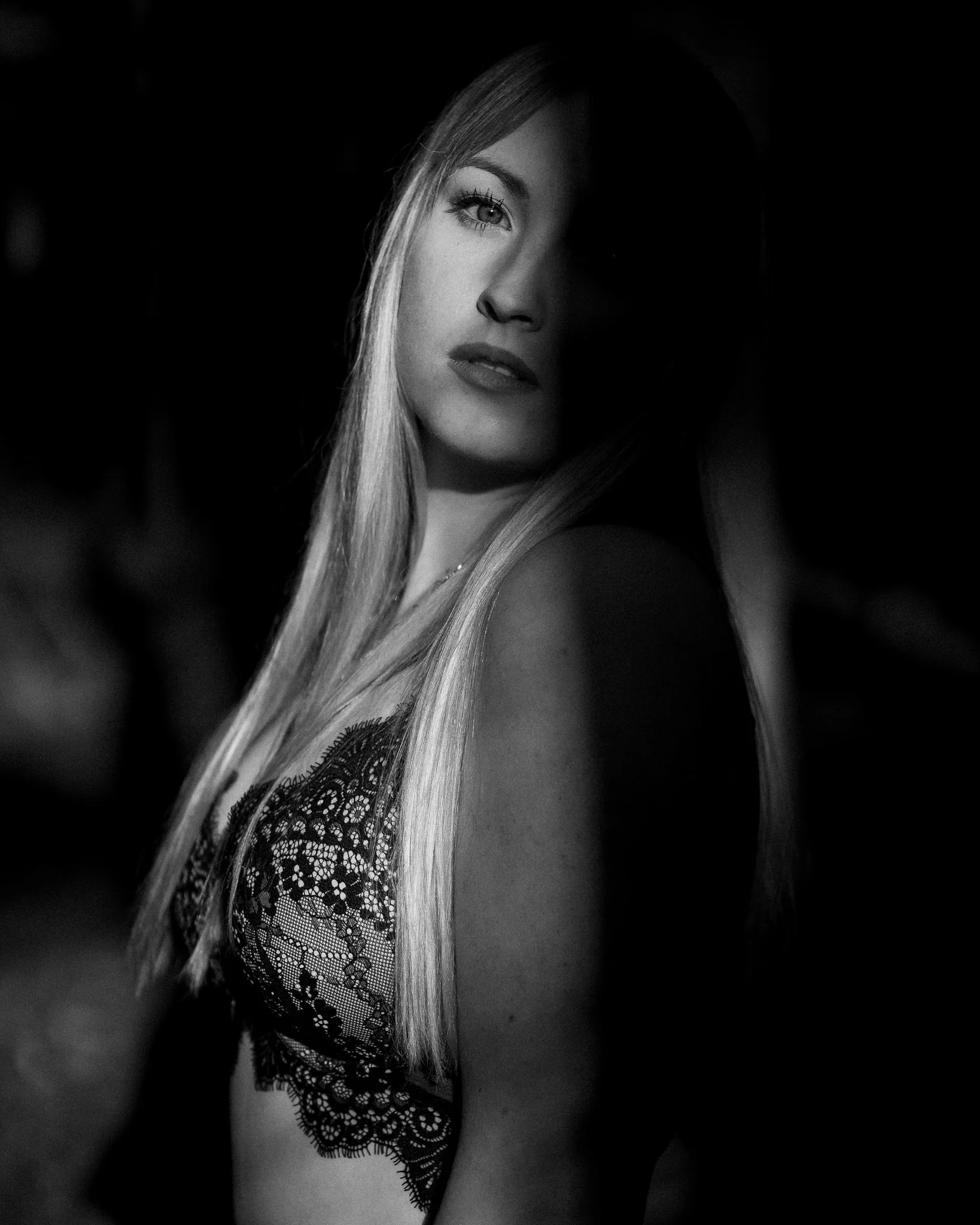 Black and white portrait of a woman with long blonde hair, one eye visible, wearing a lace top, dark background.