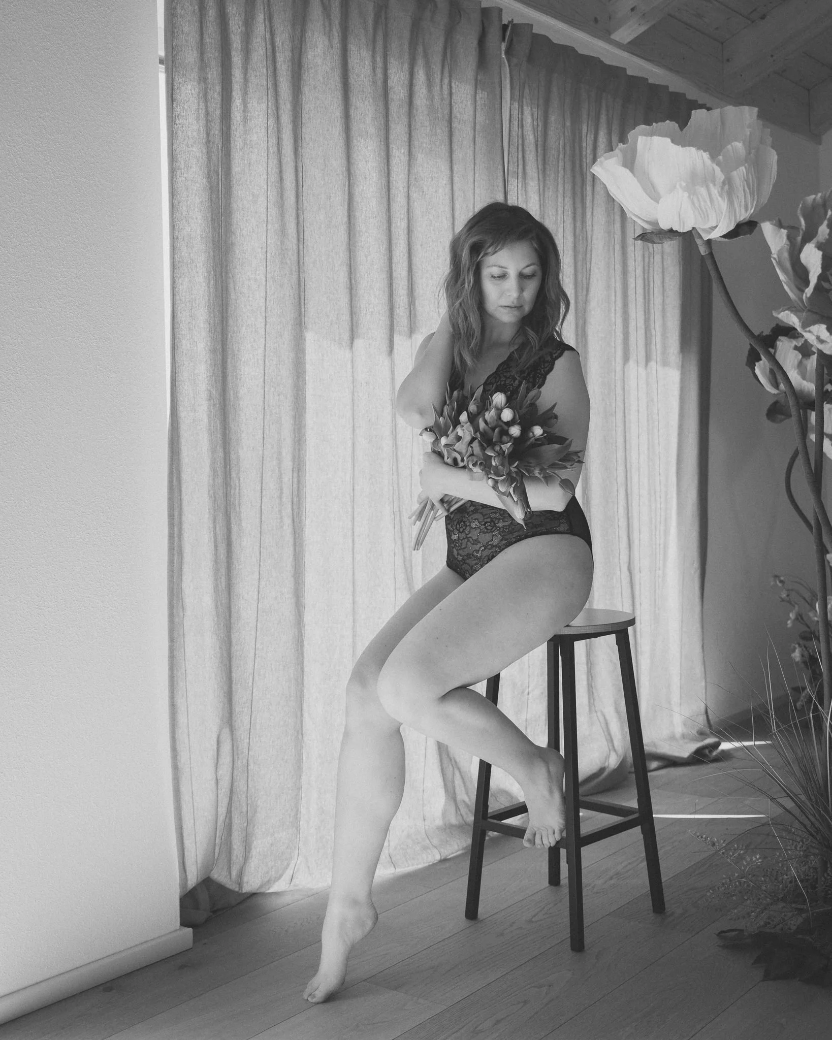 A woman with wavy hair wearing lace underwear, sitting on a stool with one leg raised and resting on the floor, holding a bouquet of flowers, near a curtain and large flower decoration in a room with wooden ceiling and flooring.