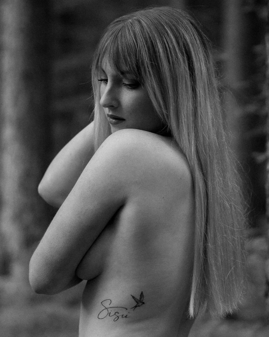 A woman with long hair and a tattoo on her side, standing sideways with her eyes closed, leaning against a tree in a natural outdoor setting, in black and white.