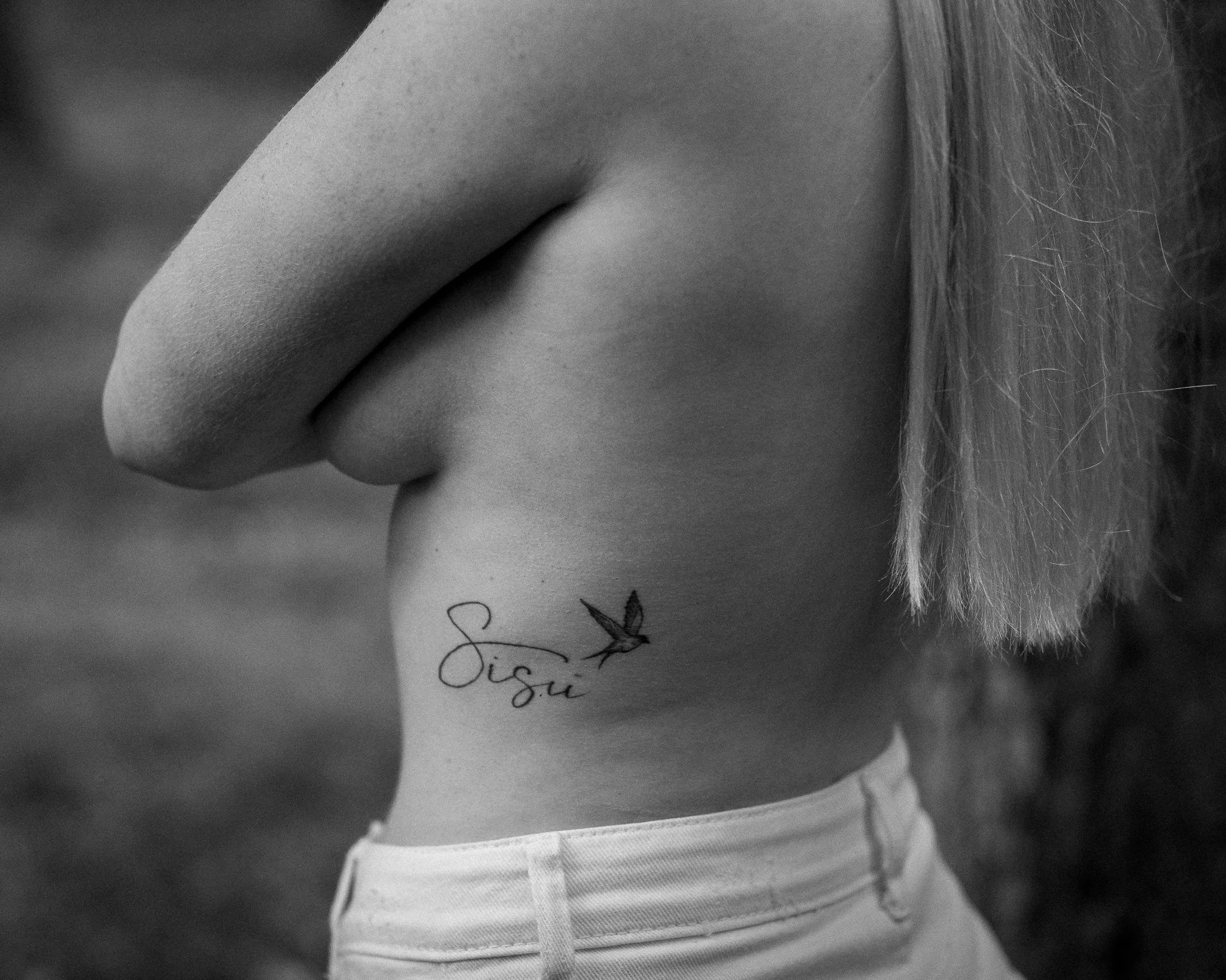 Close-up black and white photograph of a woman's face, focusing on her nose, lips, and chin, with a tattoo reading "Sio" and a hummingbird on her neck.