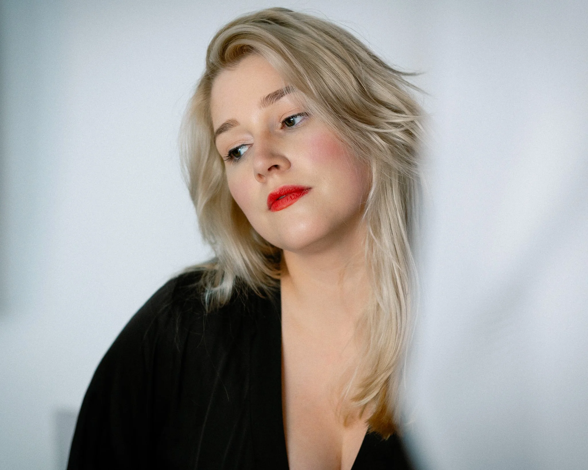 Close-up portrait of a woman with blonde, wavy hair, red lipstick, and a black top against a plain white background.