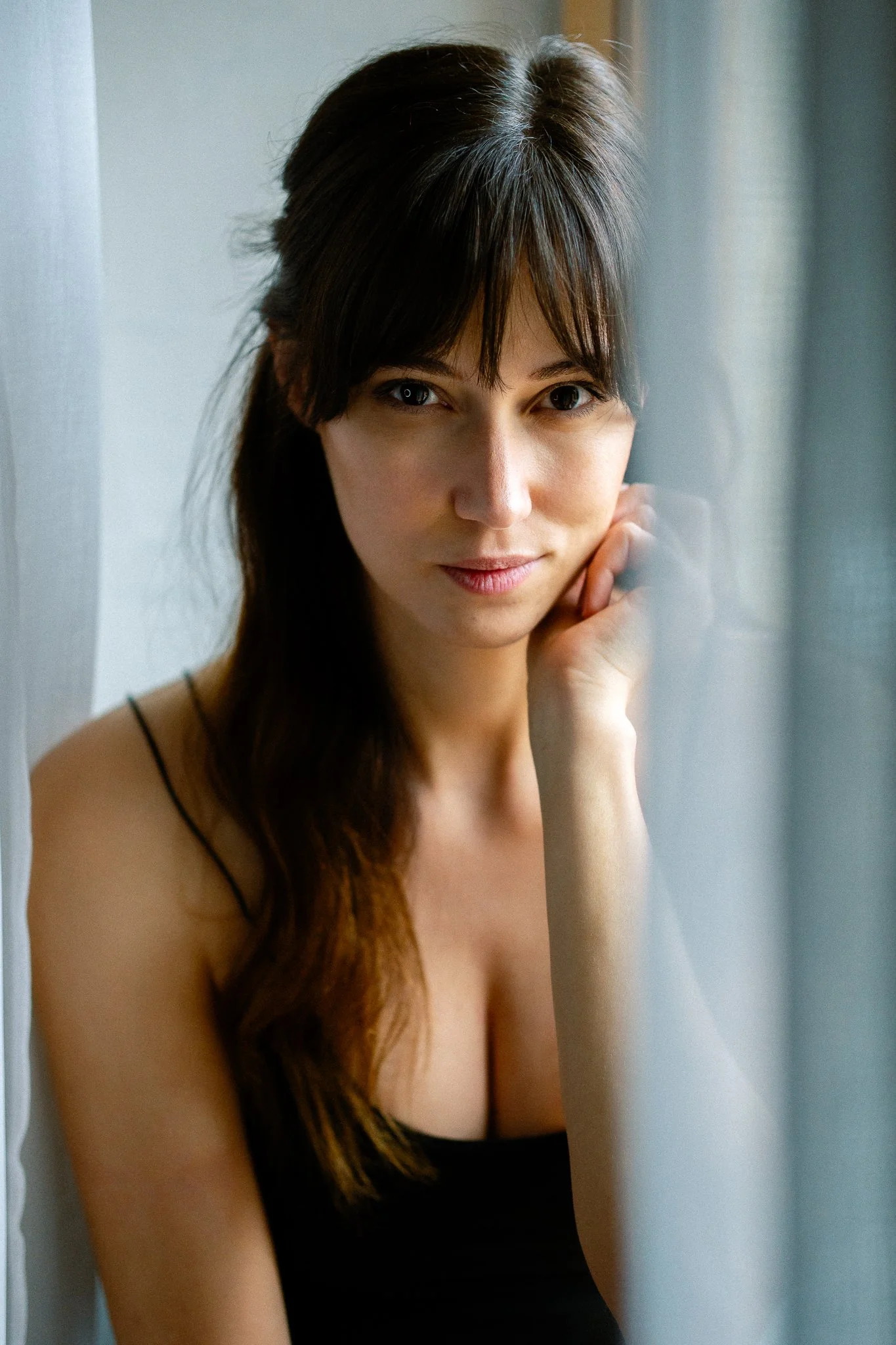 A woman with long dark hair and bangs, wearing a black spaghetti strap top, sitting beside a window with light-colored curtains, touching her face gently with one hand, looking at the camera with a subtle smile.