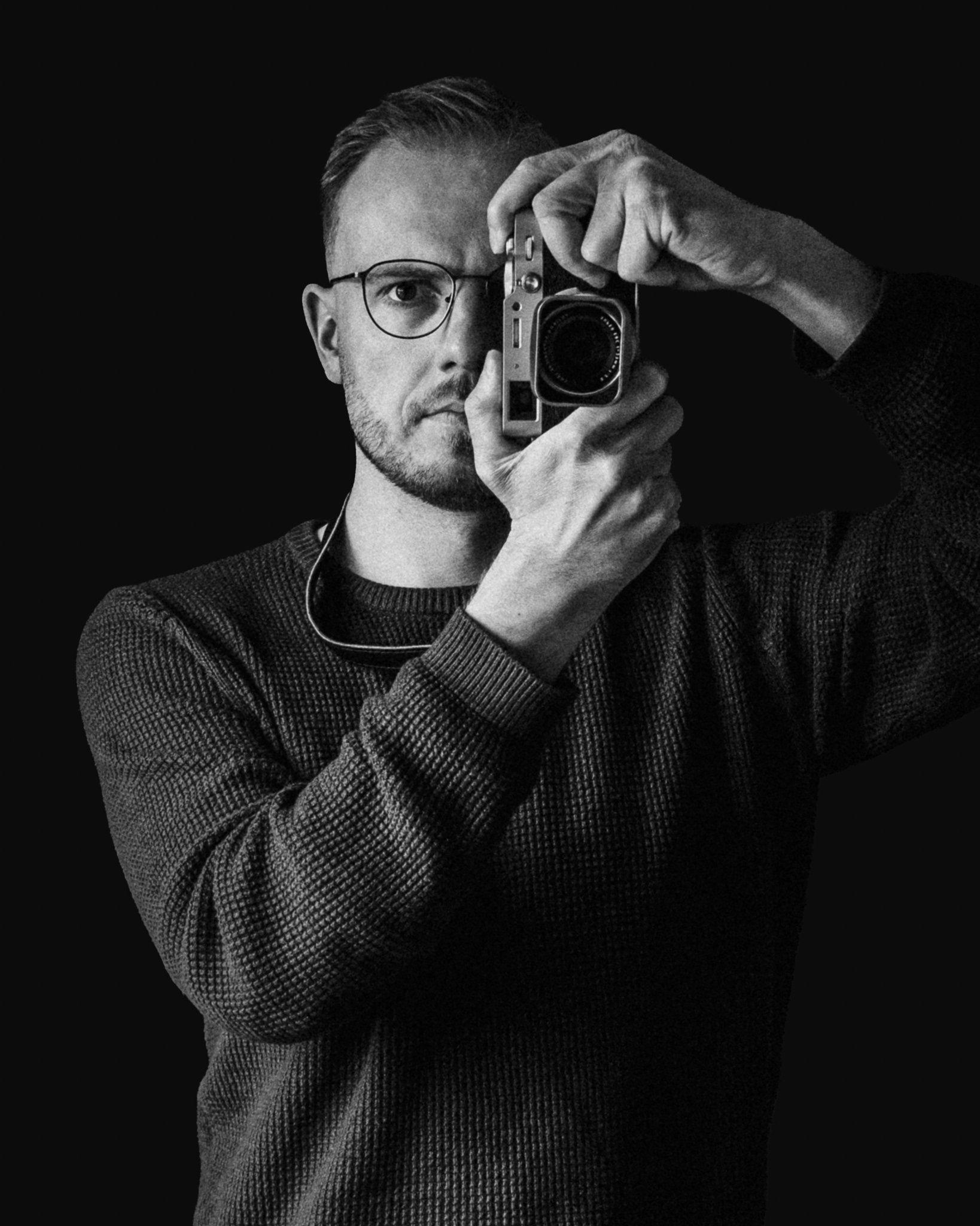 In this black and white photo, a man with glasses and short hair is holding a vintage camera up to his face, taking a photo of himself in a mirror.
