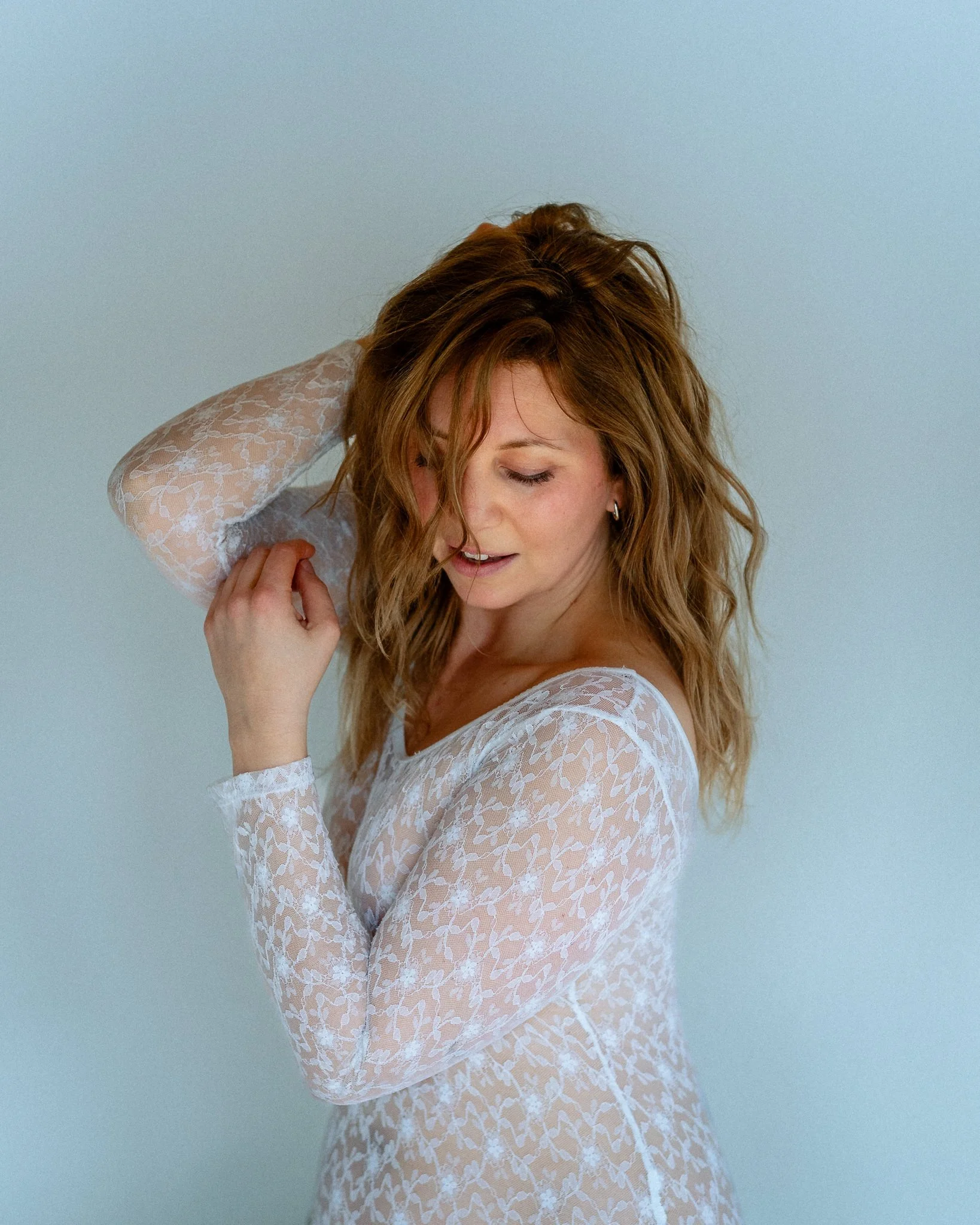 A woman with auburn hair in loose waves, wearing a white lace dress with floral patterns, posing with her eyes closed and her hand touching her hair against a plain, light-colored background.