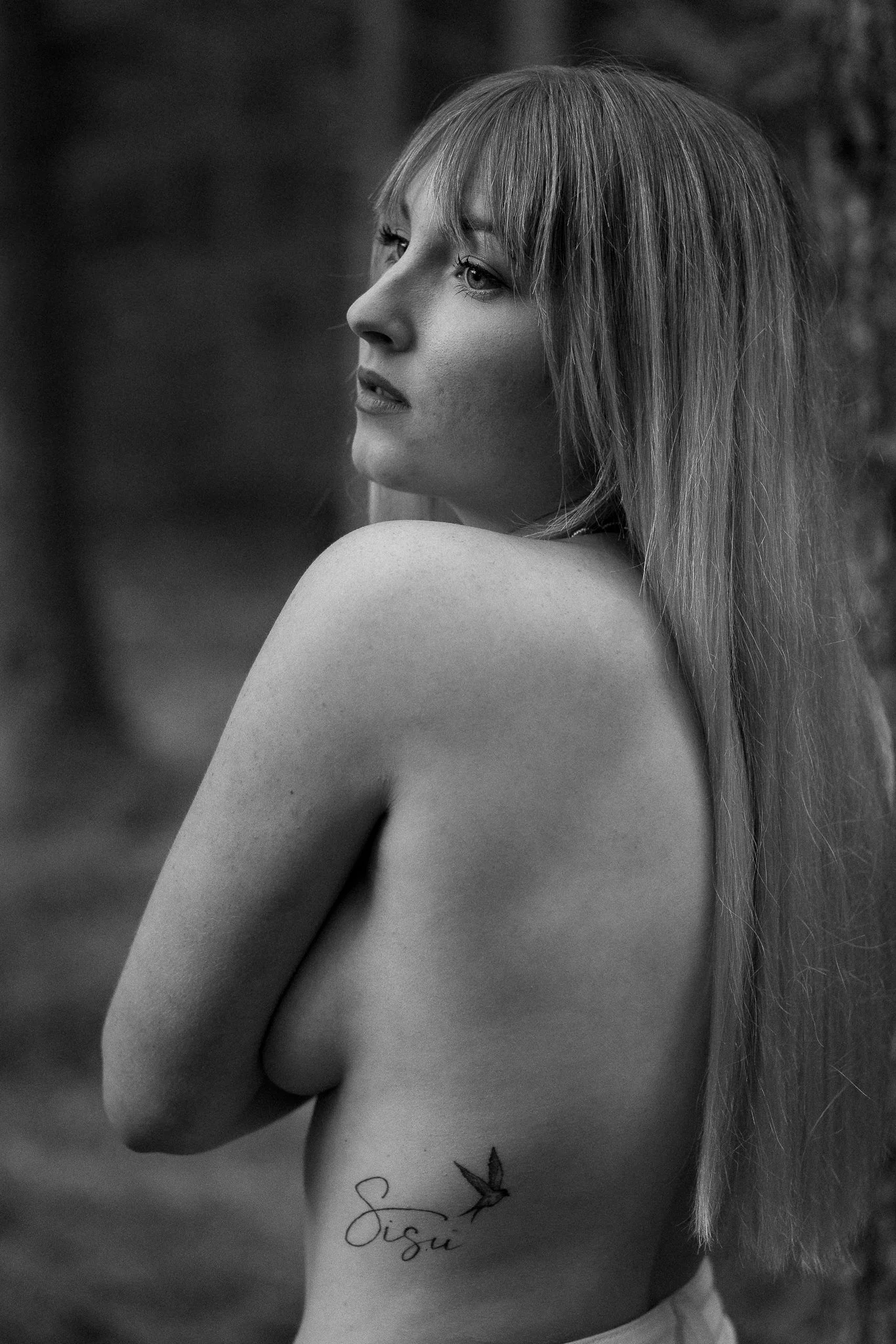 Black and white photo of a woman with long hair, looking to the side, with a tattoo of a bird and the word "Sisy" on her ribcage.