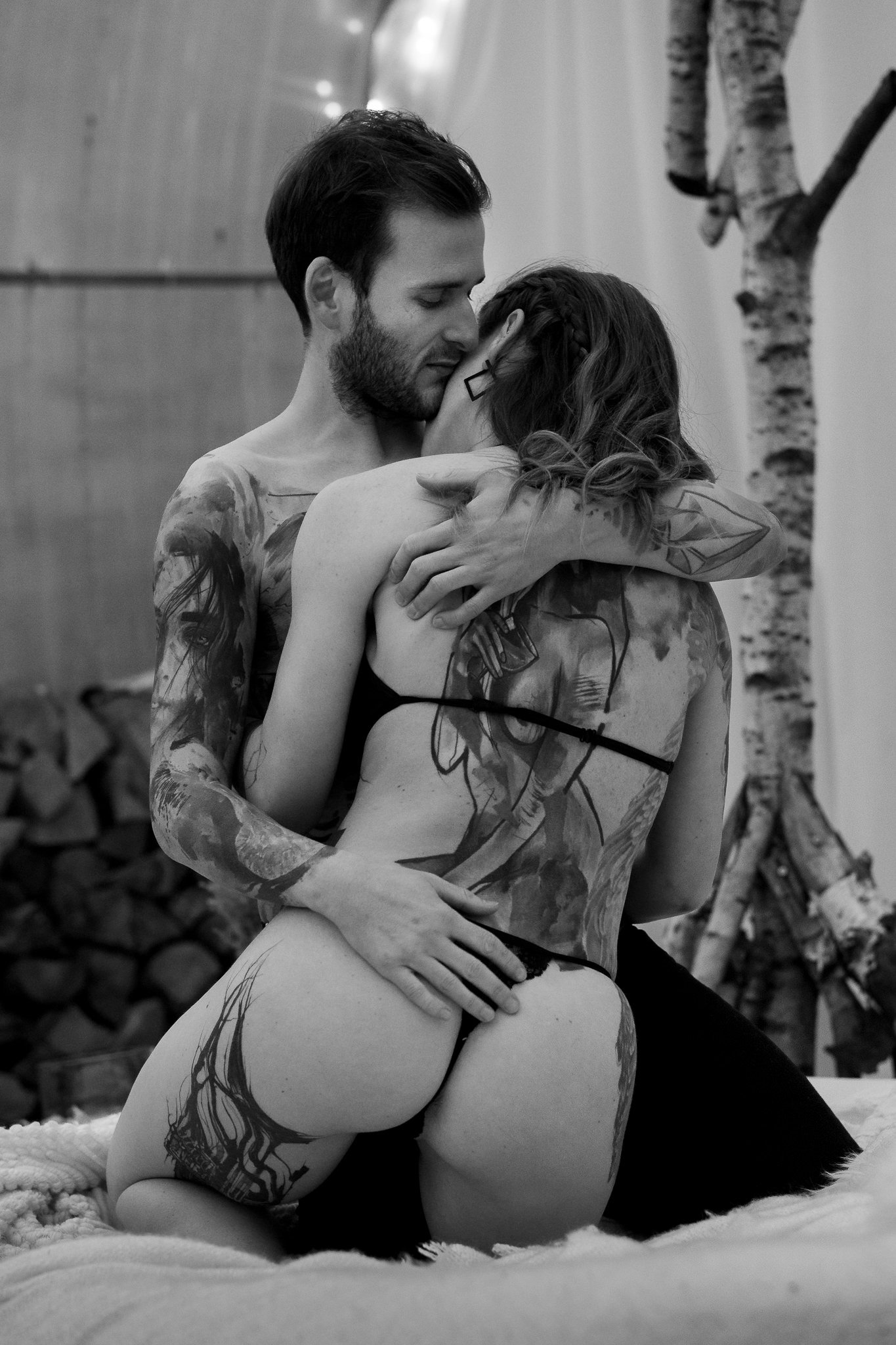 A black and white photograph of a tattooed man and woman embracing intimately. The man has dark hair and beard, and the woman has long hair braided on one side and wearing earrings. They are sitting on a soft surface with a forest of trees in the background.