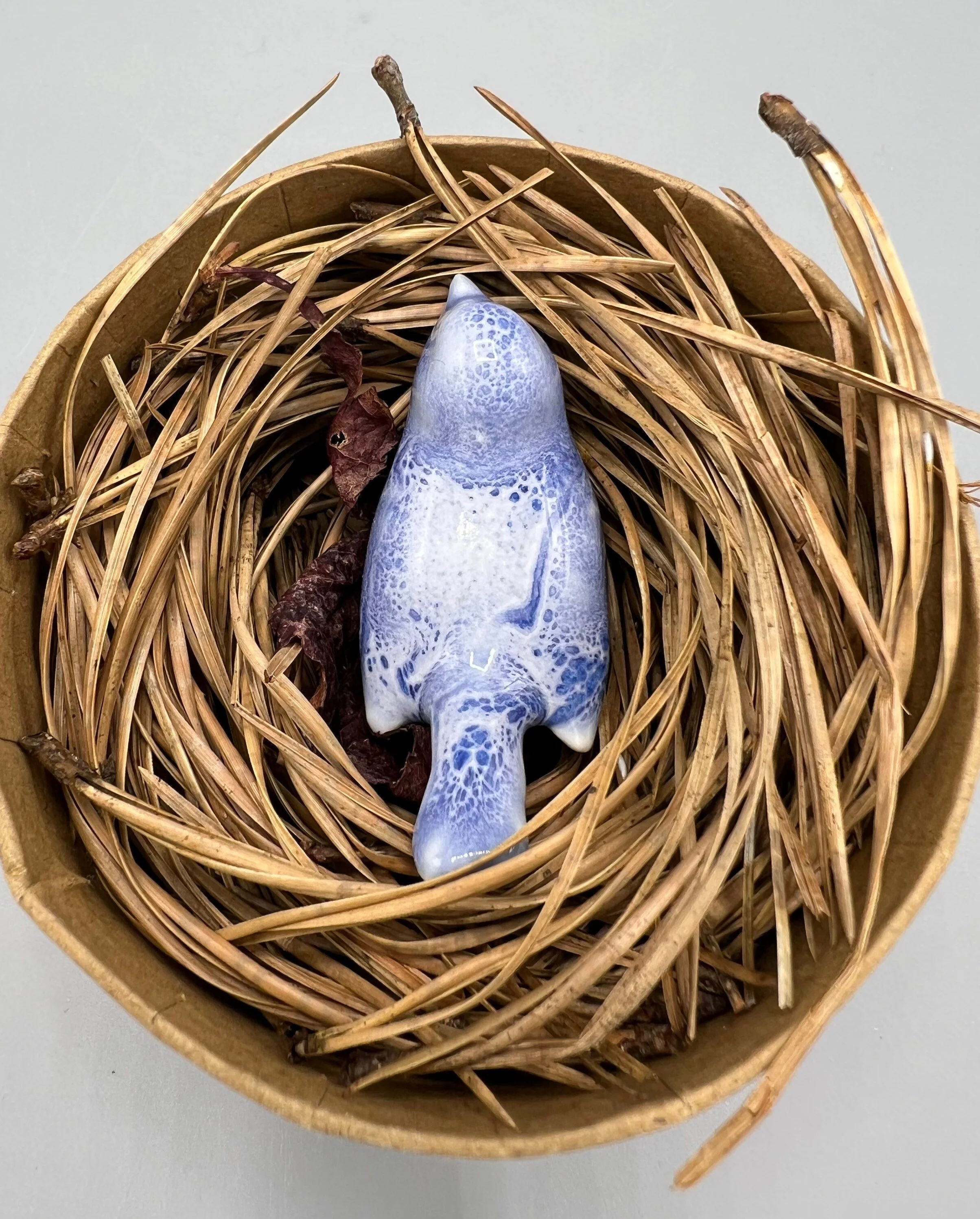 Small ceramic decorative bird, placed on a nest of dried leaves and twigs inside a round box.