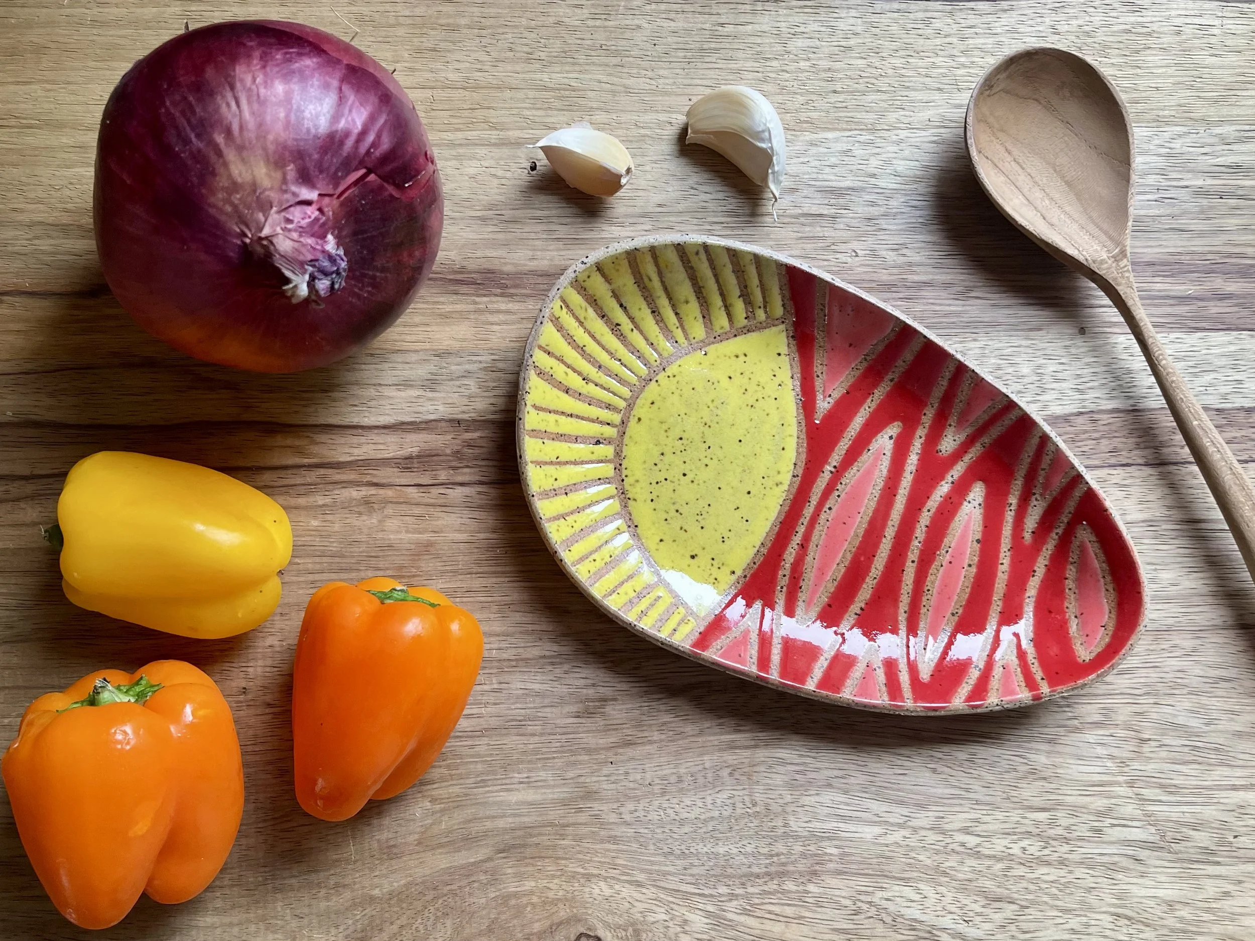A wooden table with a red onion, a yellow bell pepper, two orange bell peppers, two garlic cloves, a small wooden spoon, and a colorful ceramic dish containing olive oil and a red sauce or condiment.