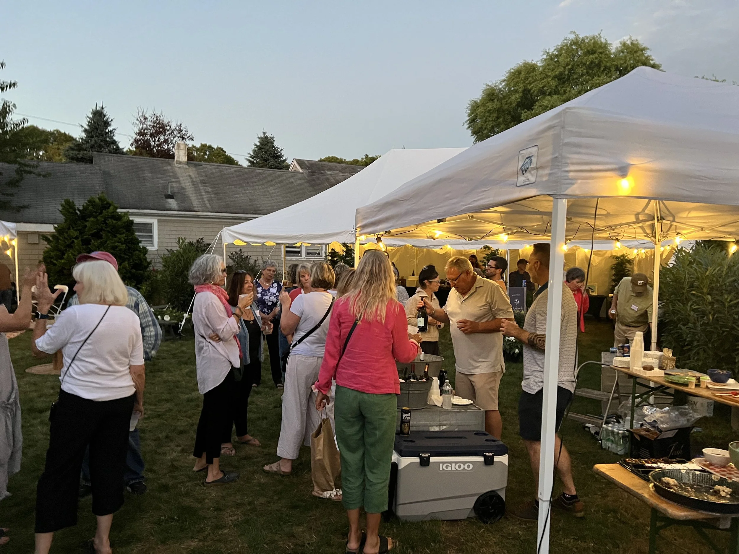 People gathered at an outdoor evening reception under canopy tent, chatting, and enjoying food and drinks in a garden setting.