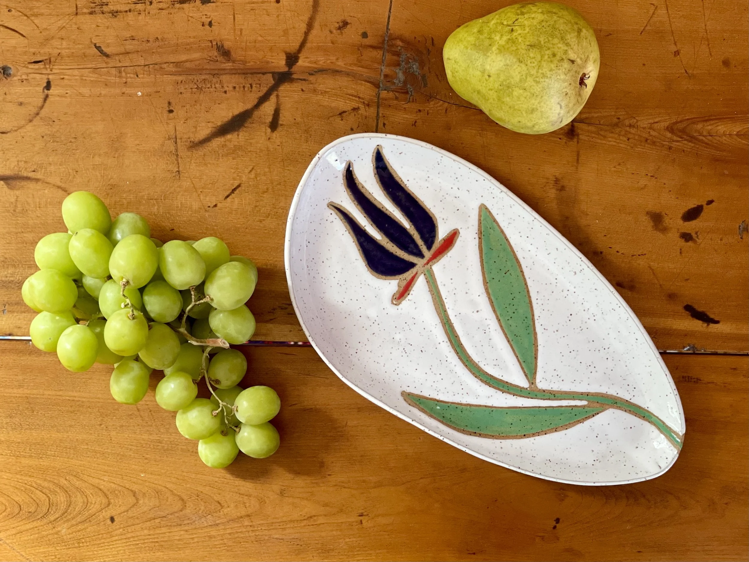 A bunch of green grapes, a pear, a decorative oval dish with a floral design, all placed on a wooden surface.