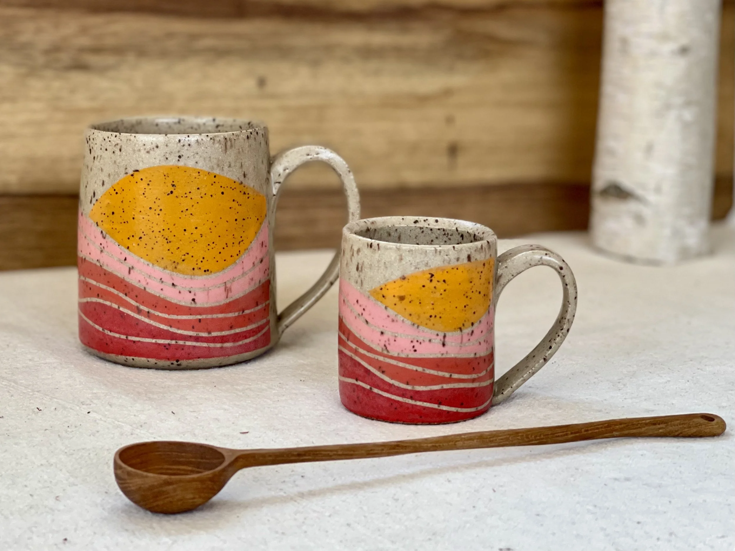 Two ceramic mugs with abstract pink, red, and orange art, placed on a white surface, alongside a wooden spoon, with a wooden background and a white log in the background.
