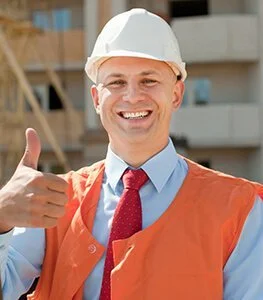 Goed geklede bouwvakker met een witte helm en oranje veiligheidsjack die glimlacht en duimen omhoog doet op een bouwplaats.
