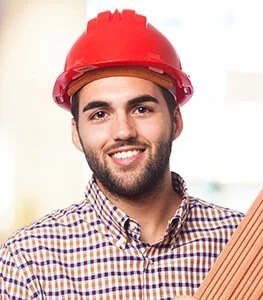 Man met een rode bouwhelm en een geruit overhemd, lachend, binnen.