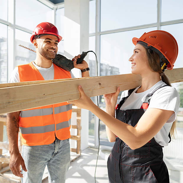 Twee bouwvakkers, een man en een vrouw, die samen een houten balk vasthouden in een bouwplaats met grote ramen en veel natuurlijk licht, beiden dragen oranje veiligheidshelmen.