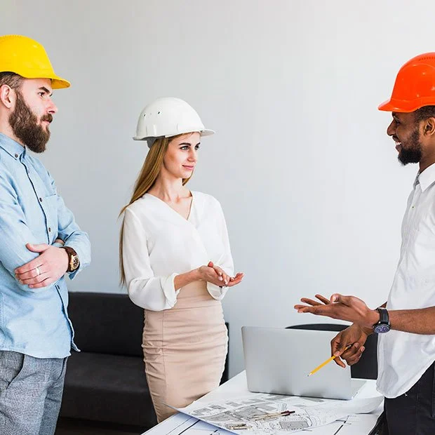 Drie mensen in een kantoor, allemaal met veiligheidshelmen, die een gesprek voeren.