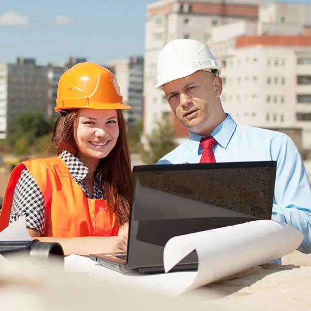 Twee bouwvakkers, een vrouw met een oranje helm en een man met een witte helm, bekijken documenten en een laptop op een bouwplaats met gebouwen op de achtergrond.