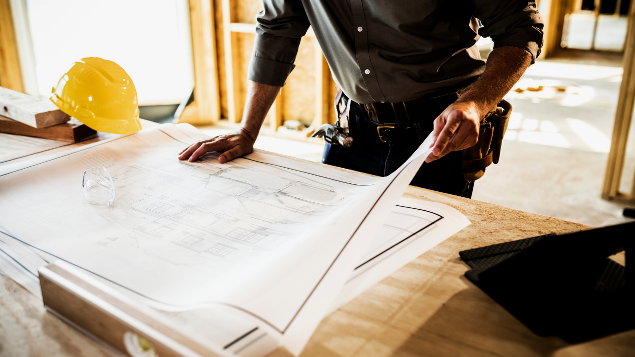 Mannen die bouwplannen bekijkt op een bouwplaats, met een gele bouwhelm op de tafel.