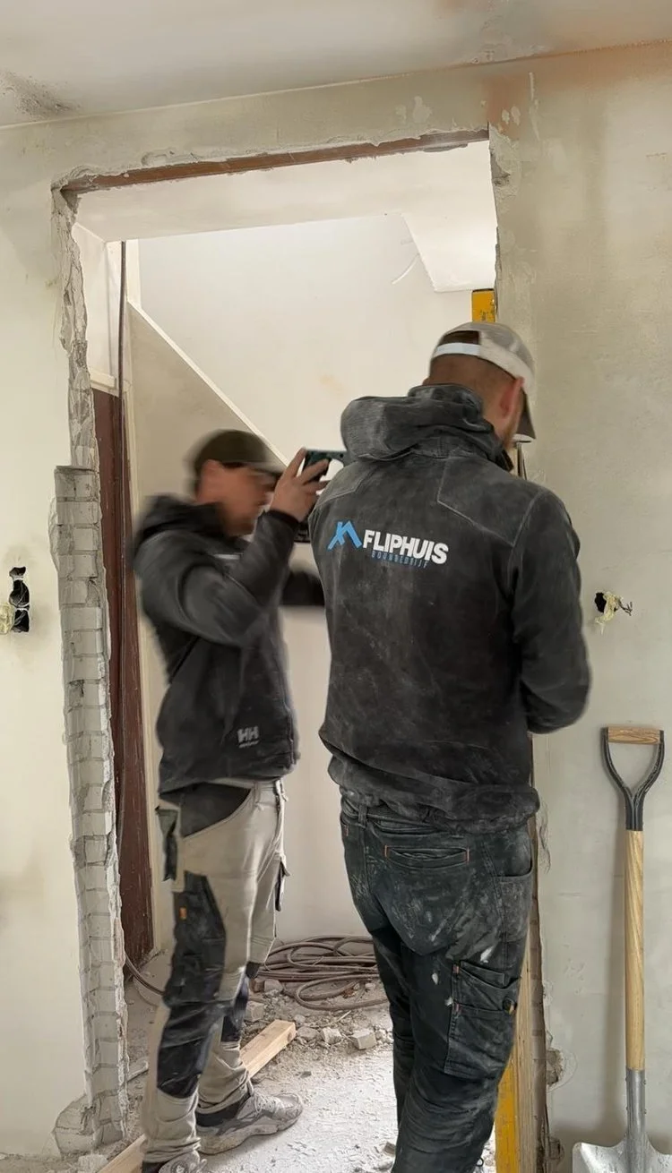 Twee vakmensen werken aan een binnenmuur, één met een waterpas en de ander met een tikcomputer, in een verbouwingsproject.