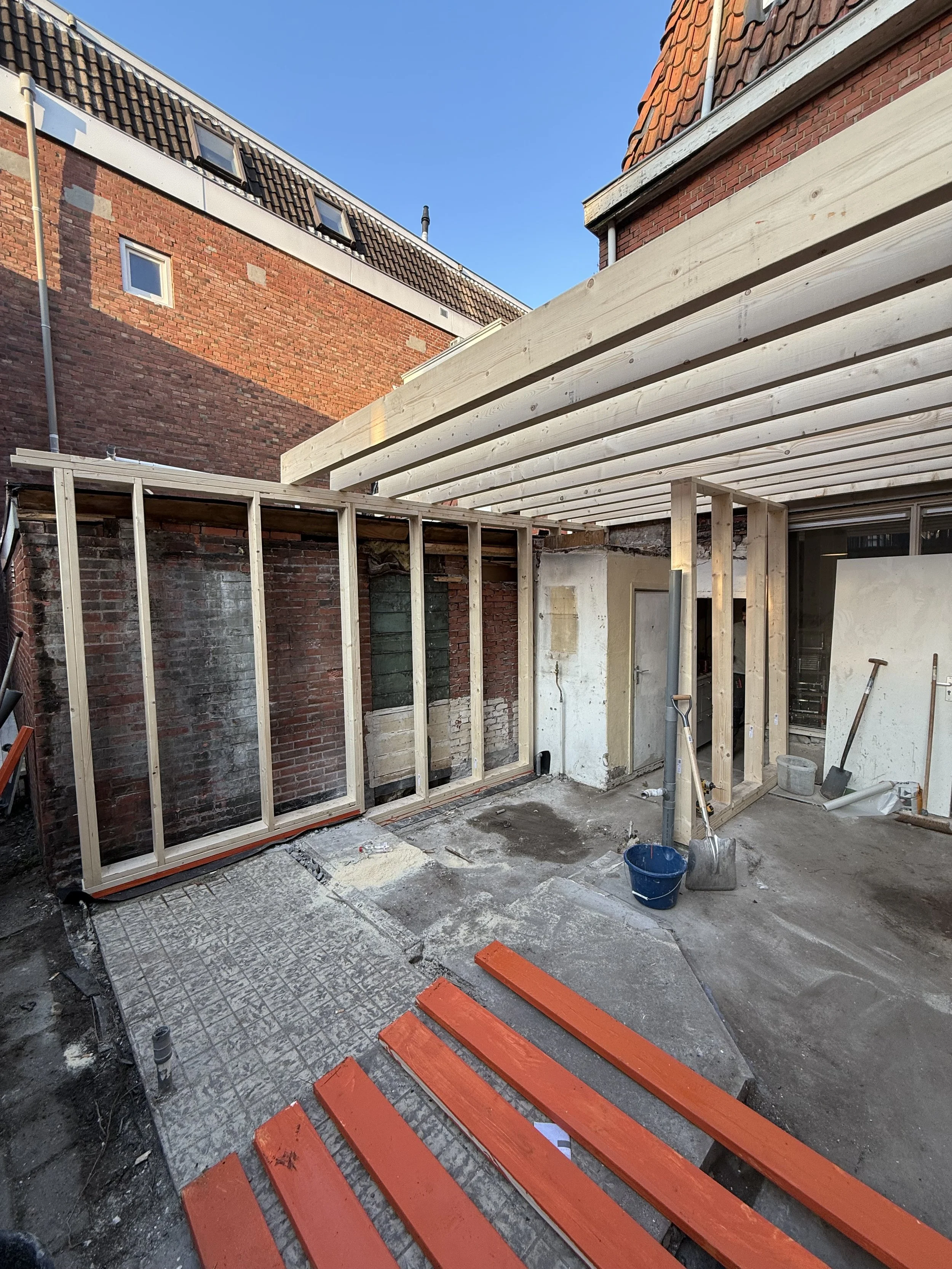 Bouwplaats met houten structuur, afgebroken muur, en verfvlammen met oranje planken voor projectuitbreiding.