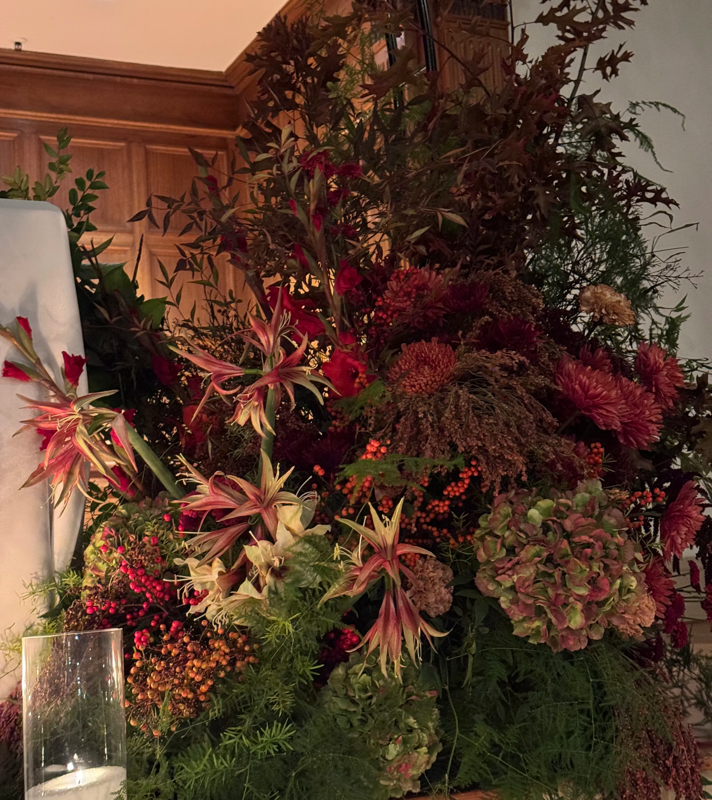 A large floral arrangement of red, pink, and purple flowers and green foliage, set against a wooden wall and illuminated by soft lighting.