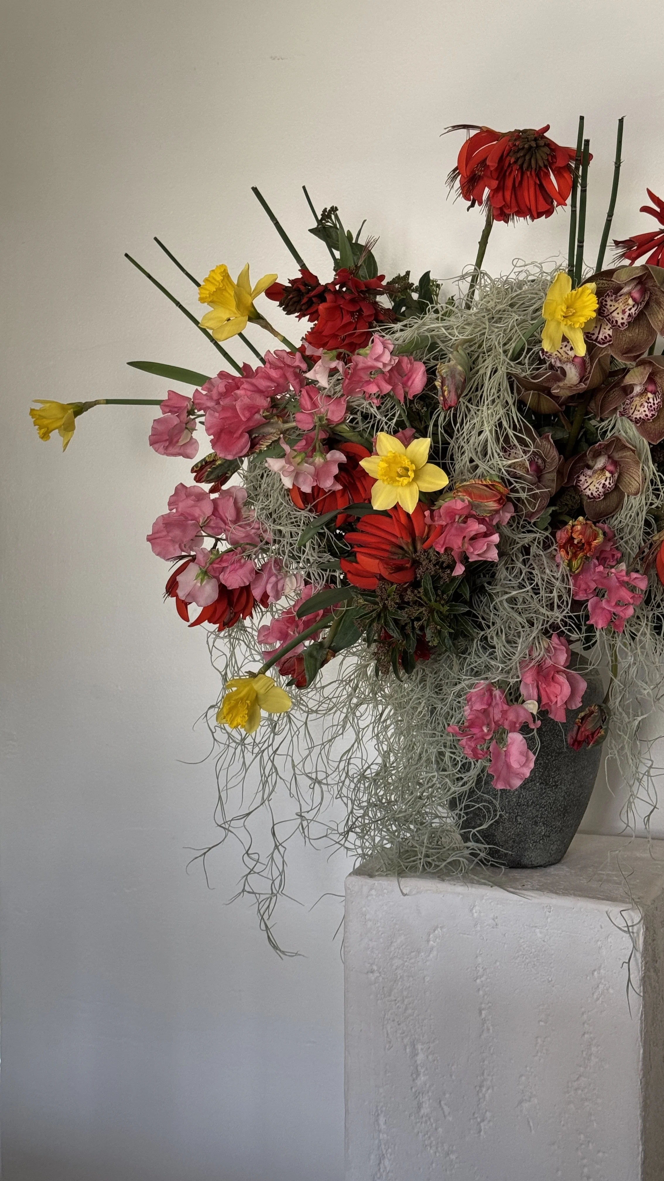 A large bouquet of various colorful flowers, including pink, yellow, red, and purple, in a dark textured vase on a white pedestal against a plain white wall.