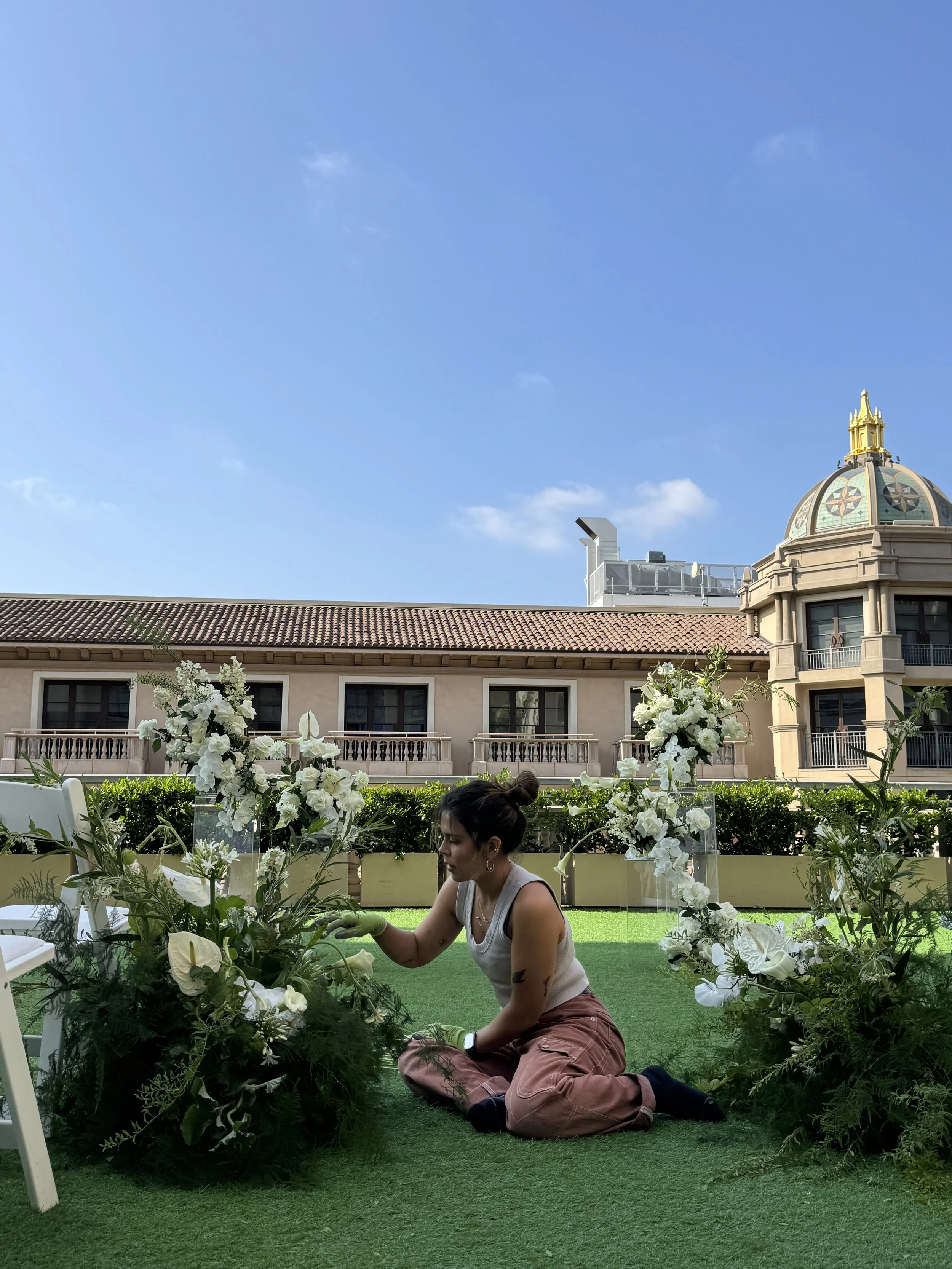A woman with dark hair in a bun, wearing a white tank top, pink pants, and green gloves, is kneeling on green artificial grass, arranging white flowers with green foliage in an outdoor setting. Behind her is a building with a tiled roof and a decorative dome with a gold finial, under a bright blue sky with a few small clouds.