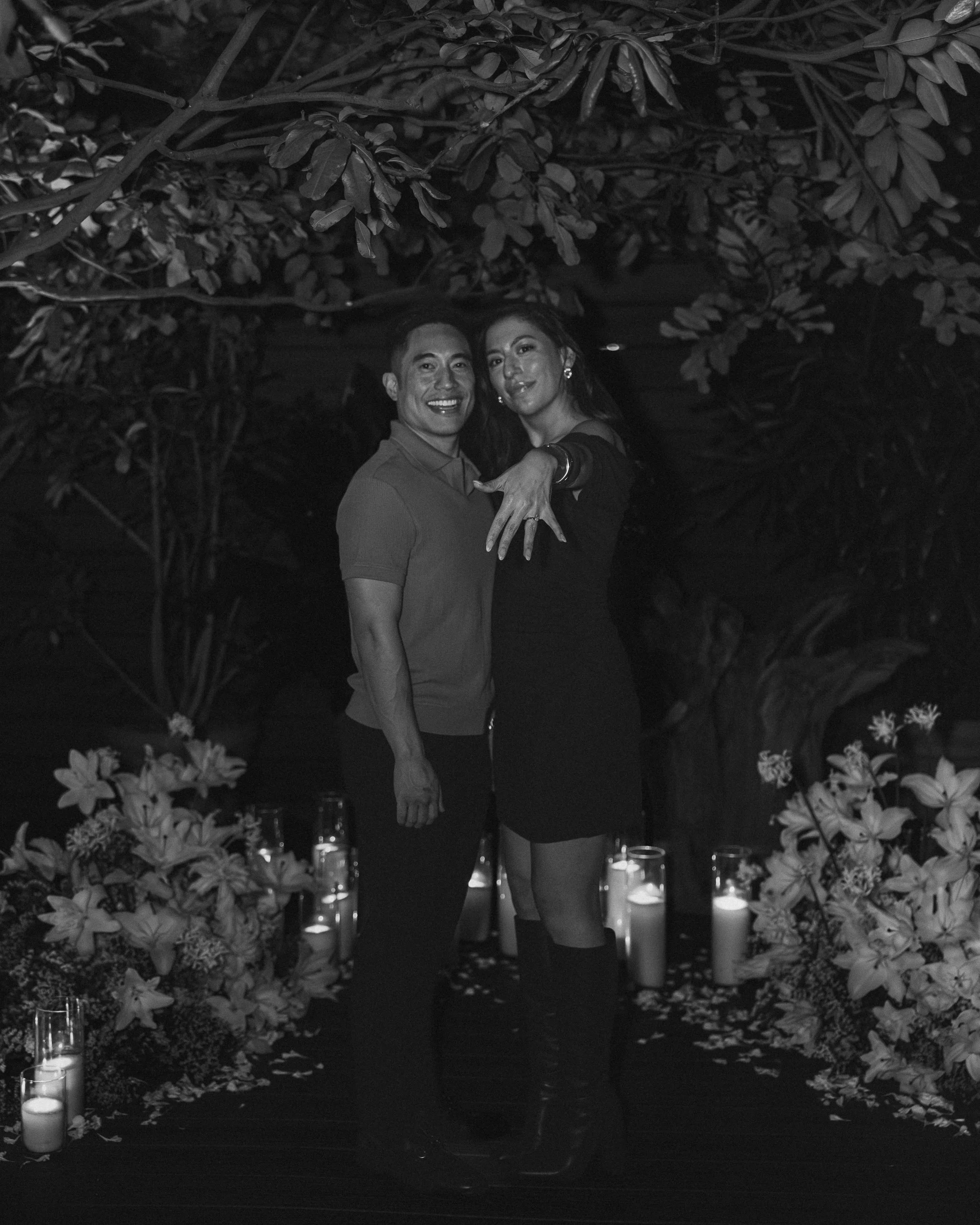 A couple embraces under a canopy of leaves and flowers at night, surrounded by lit candles and flowers on the floor, with two glasses on the ground.