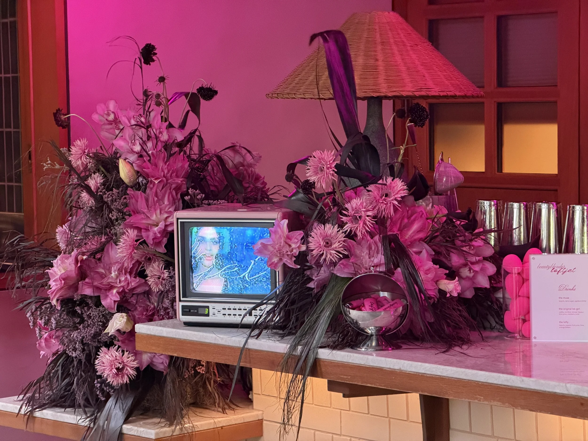 A floral display with pink flowers and greenery surrounds a small monitor on a table. A wicker table lamp is behind the flowers, and a sign to the right displays a drink menu with pink and white colors. The background includes a pink-tinted wall and a wooden window frame.