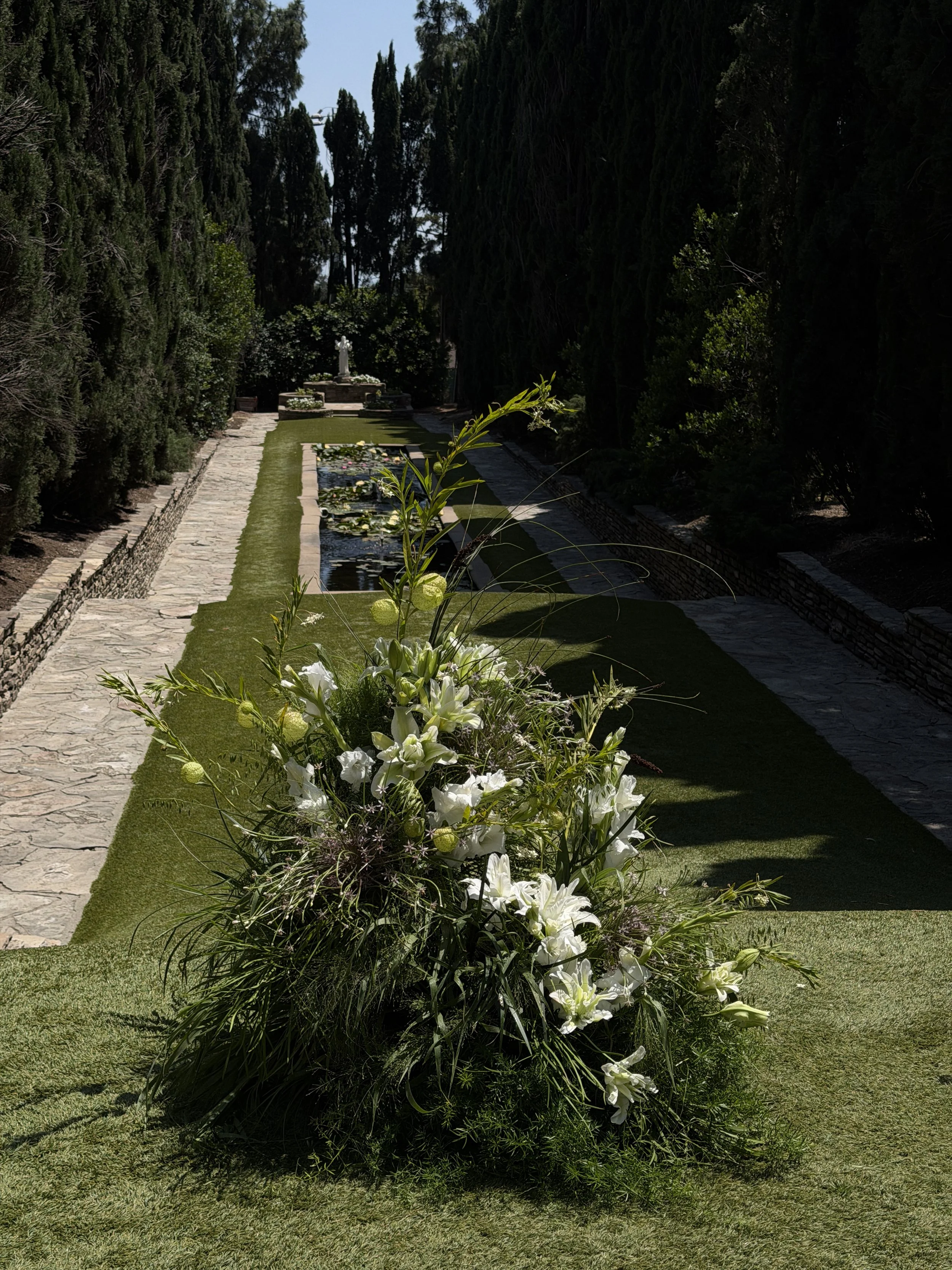 A large floral arrangement with white lilies and greenery in a garden with a stone pathway, trees, and a statue in the background.
