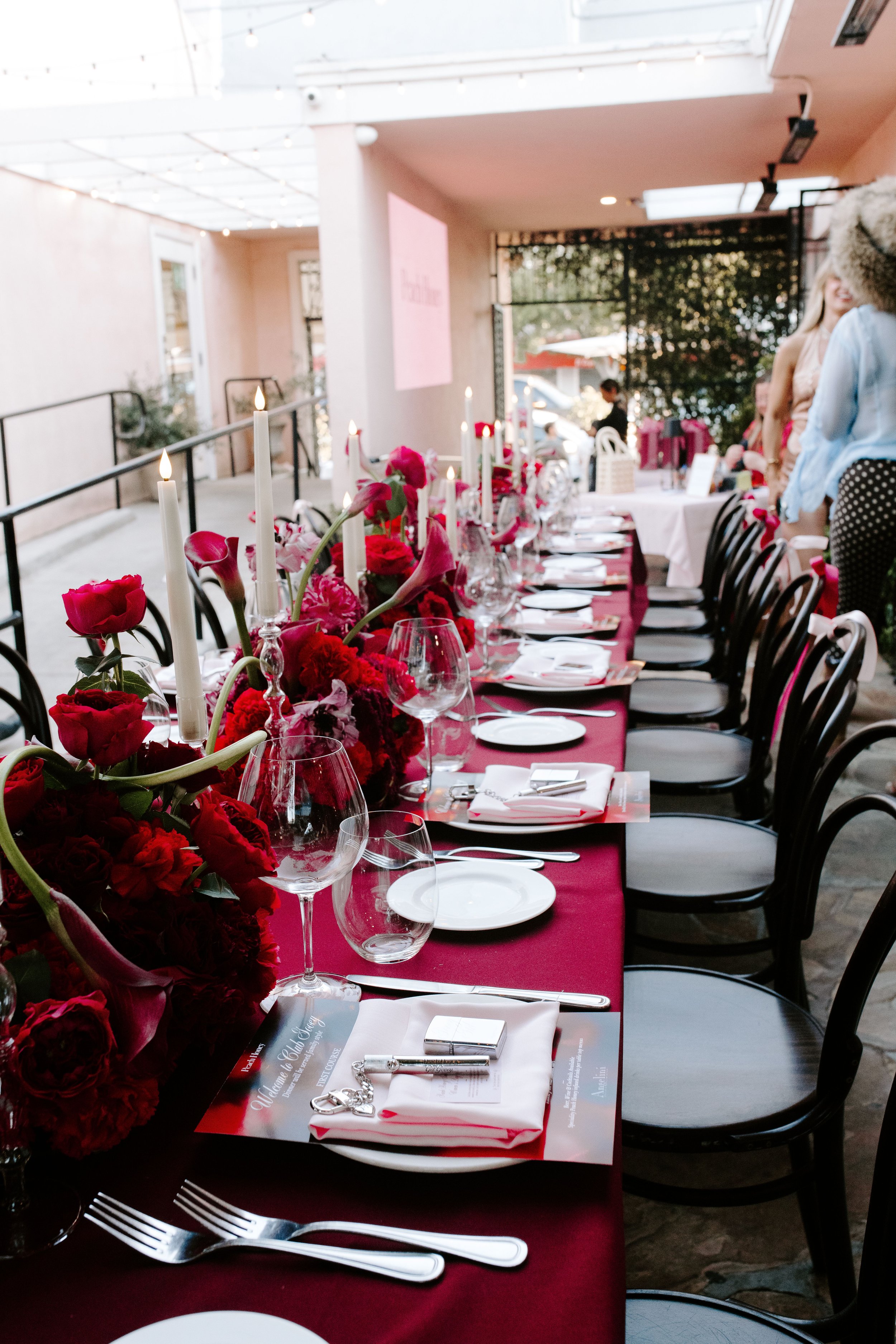 A long dining table set for a formal event with a red tablecloth, floral arrangement of red and pink flowers, white candles, wine glasses, white plates, and cutlery. The setting is indoors with large windows and a high ceiling with white string lights. There are people in the background, some standing and one sitting near the window.