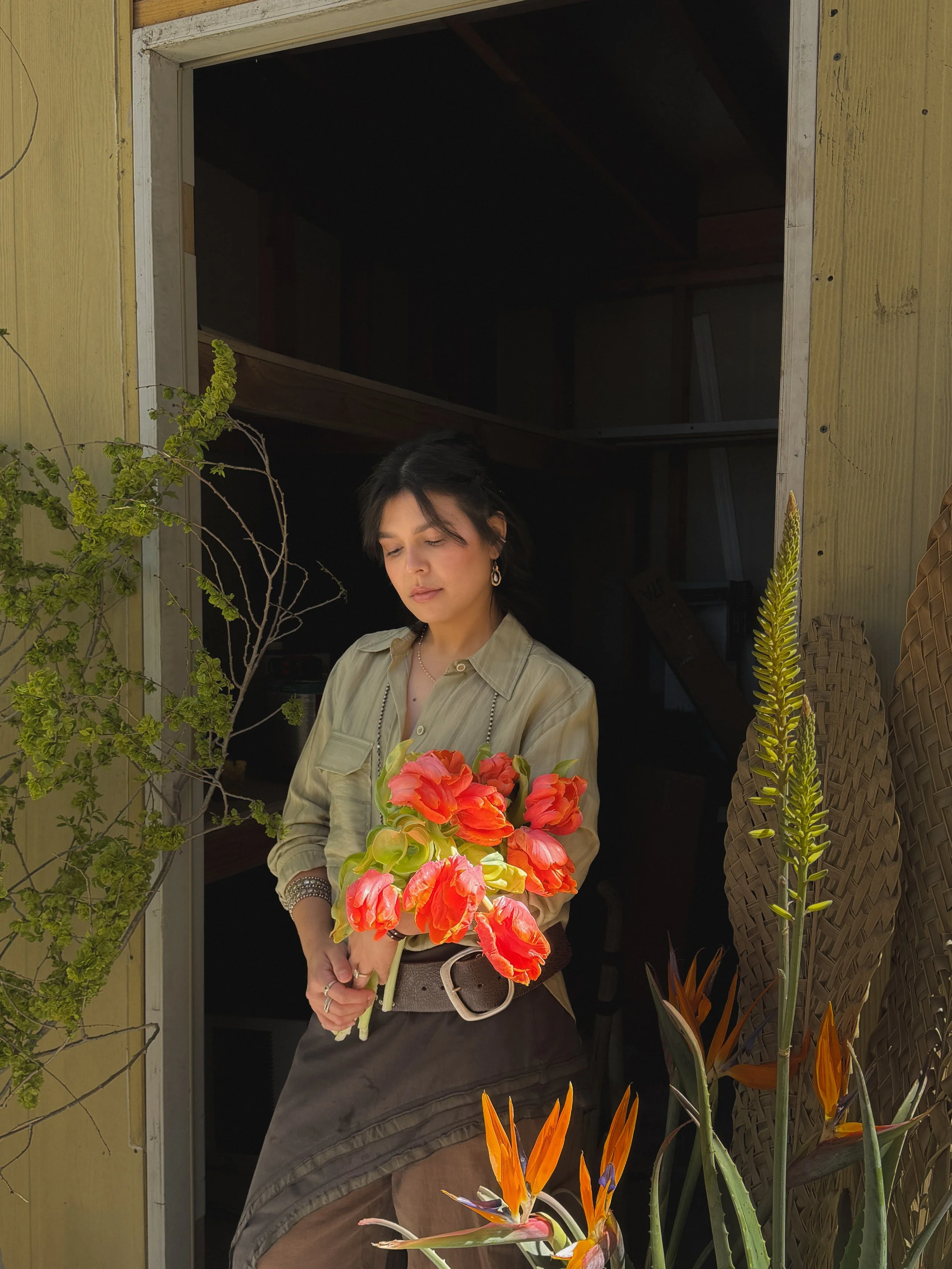 A woman standing in a doorway holding a bouquet of orange and pink flowers, surrounded by various potted plants and flowers.